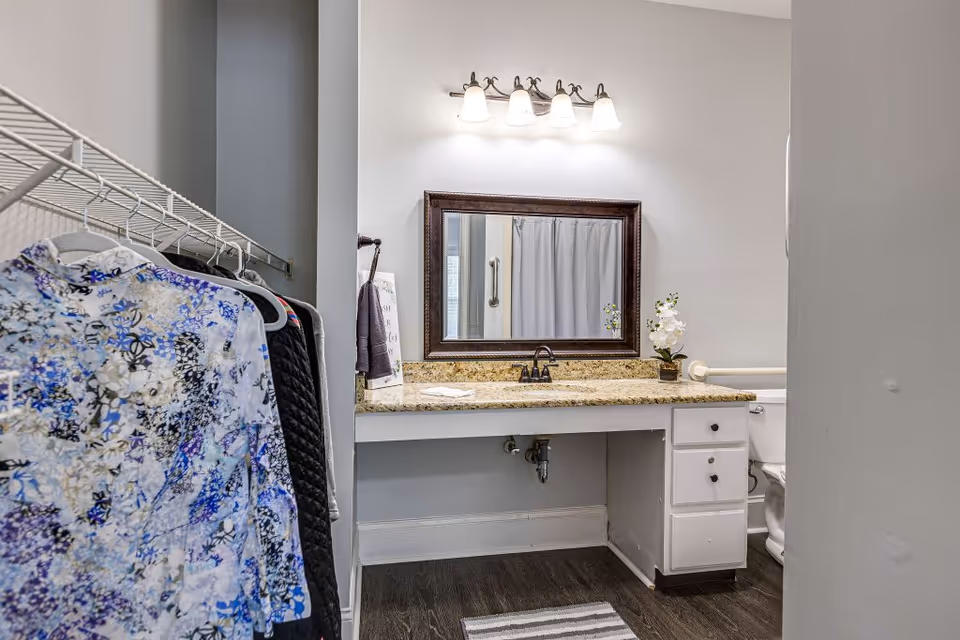A bathroom vanity area with a granite countertop, a large framed mirror, and a four-light fixture above. To the left, there is a clothing rack with several hanging garments. The bathroom has a white cabinet with drawers under the countertop and a toilet partially visible on the right side. The floor is dark wood, and a striped rug is placed in front of the vanity.