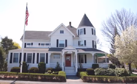 A white Victorian-style two-story house with a turret and wraparound porch, an American flag on a pole, and trimmed shrubs in front.