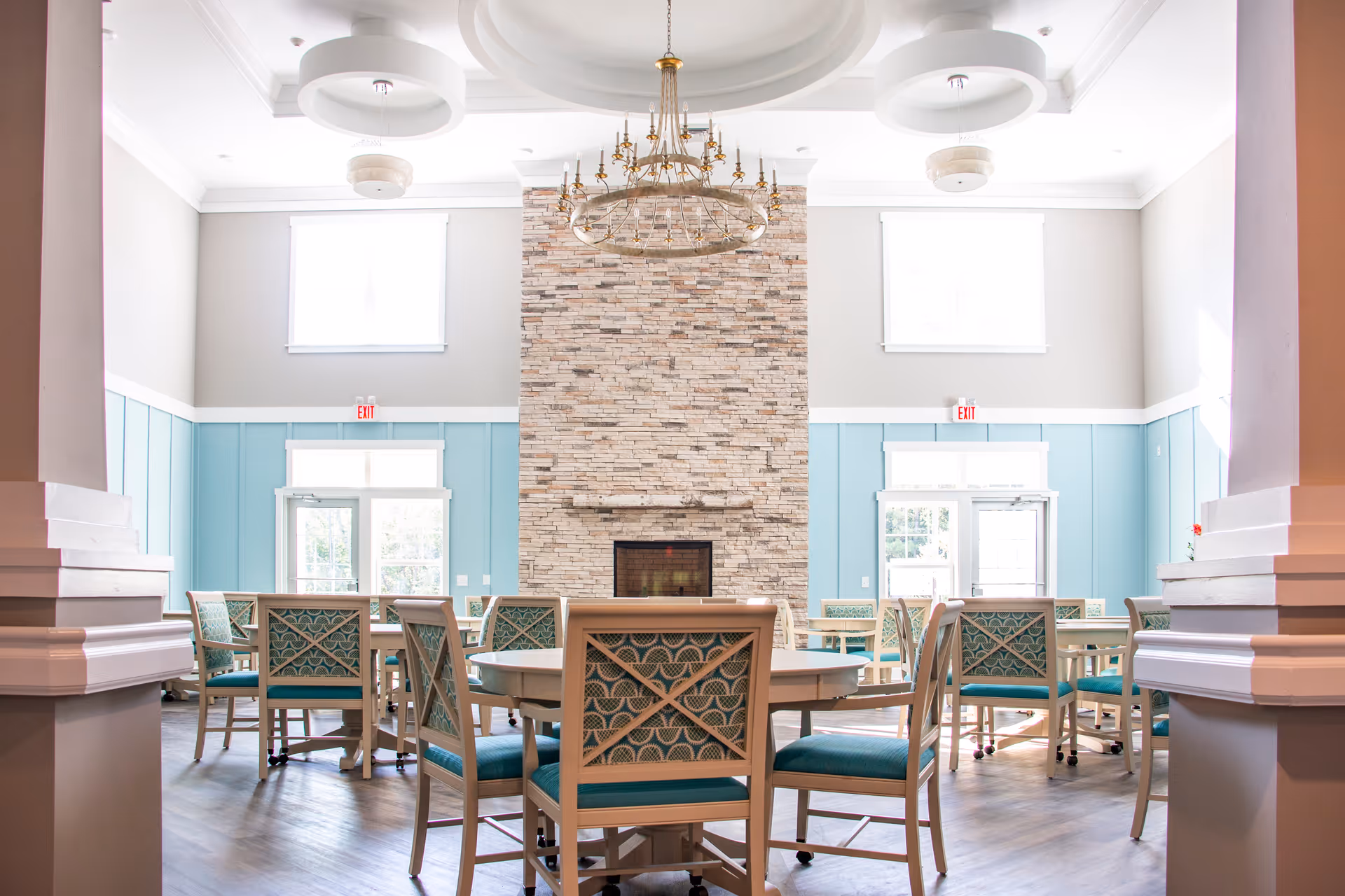 Bright communal dining room with round tables and teal-upholstered chairs facing a tall stone fireplace.