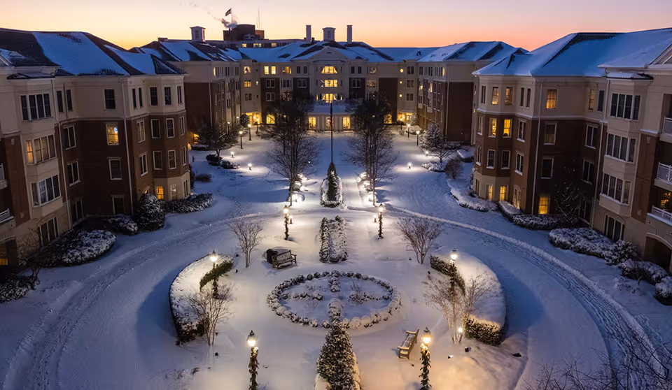 Aerial view of a senior living facility courtyard covered in snow at dusk, with lit street lamps, benches, and surrounding multi-story buildings with snow-covered roofs and lit windows.