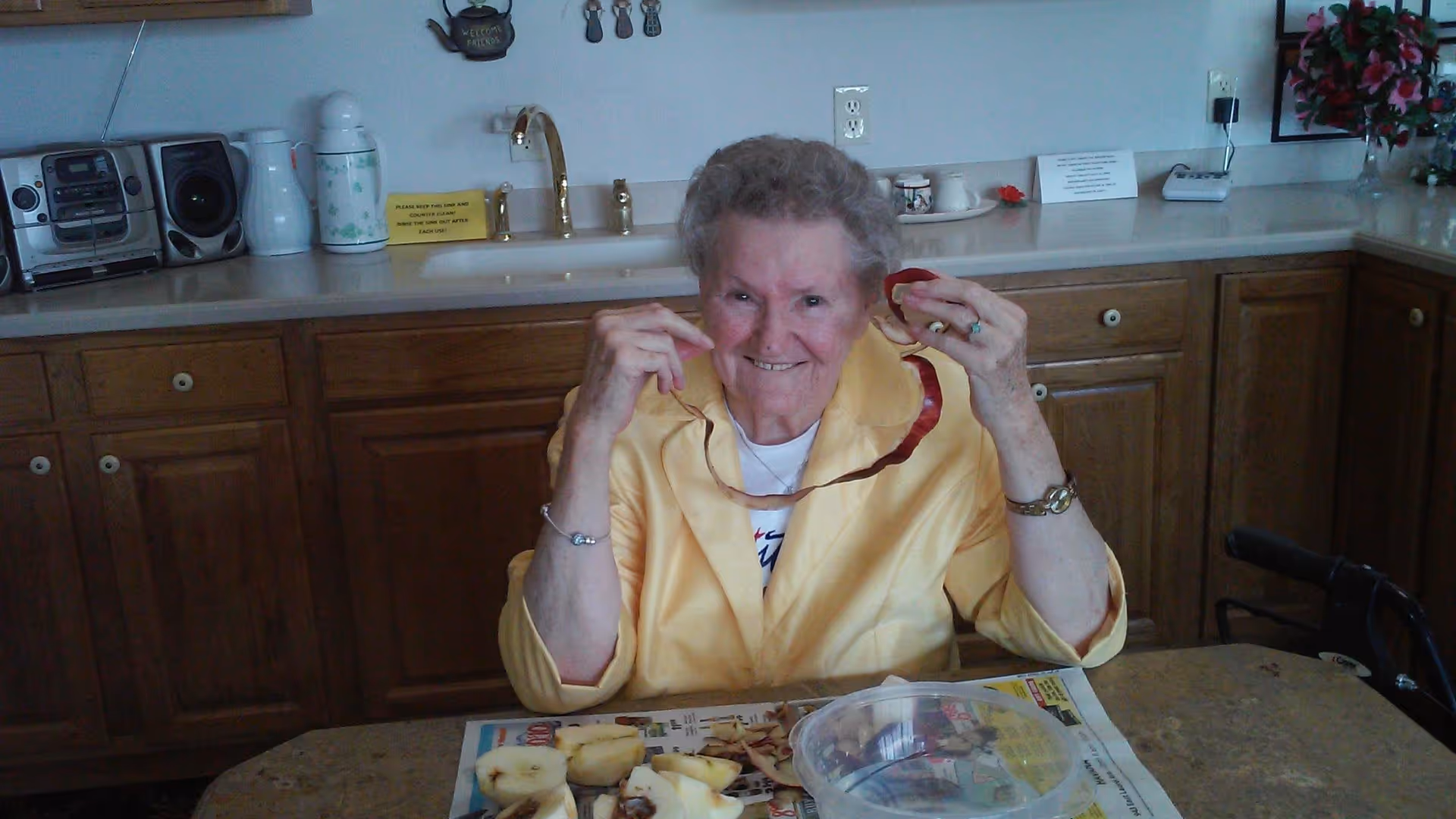An elderly woman sitting at a kitchen table peeling an apple with a smile. The kitchen has wooden cabinets, a countertop with a sink, a radio, and various kitchen items. There is a walker next to the table.