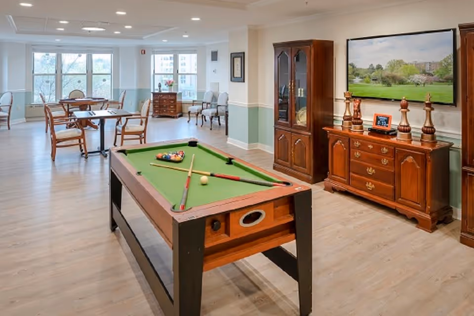 Spacious assisted-living common room with a pool table in the foreground, dining tables and chairs, wooden cabinets, and a wall-mounted TV.