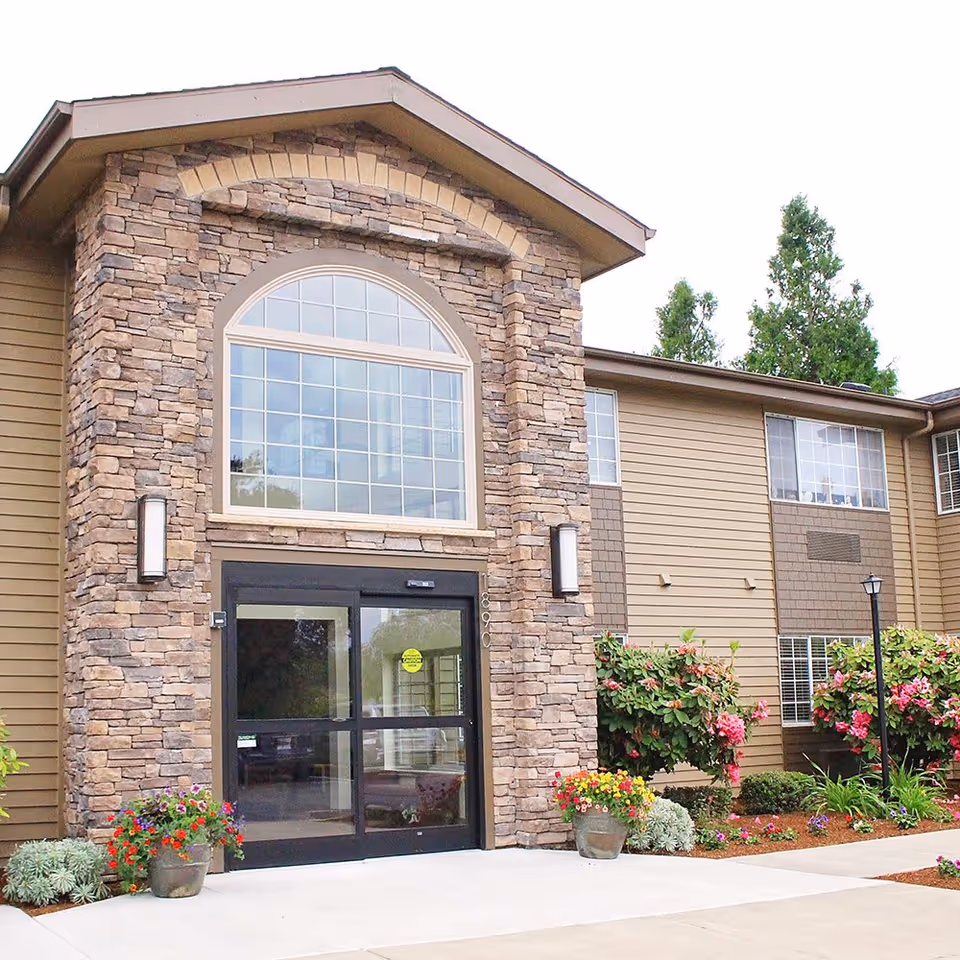 Entrance of a senior living facility with stone and wood exterior, large arched window above automatic glass doors, and landscaped flower pots and bushes on either side of the entrance.