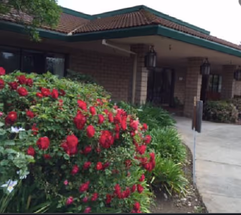 Front entrance of a brick building with a covered porch and red rose bushes in the foreground.