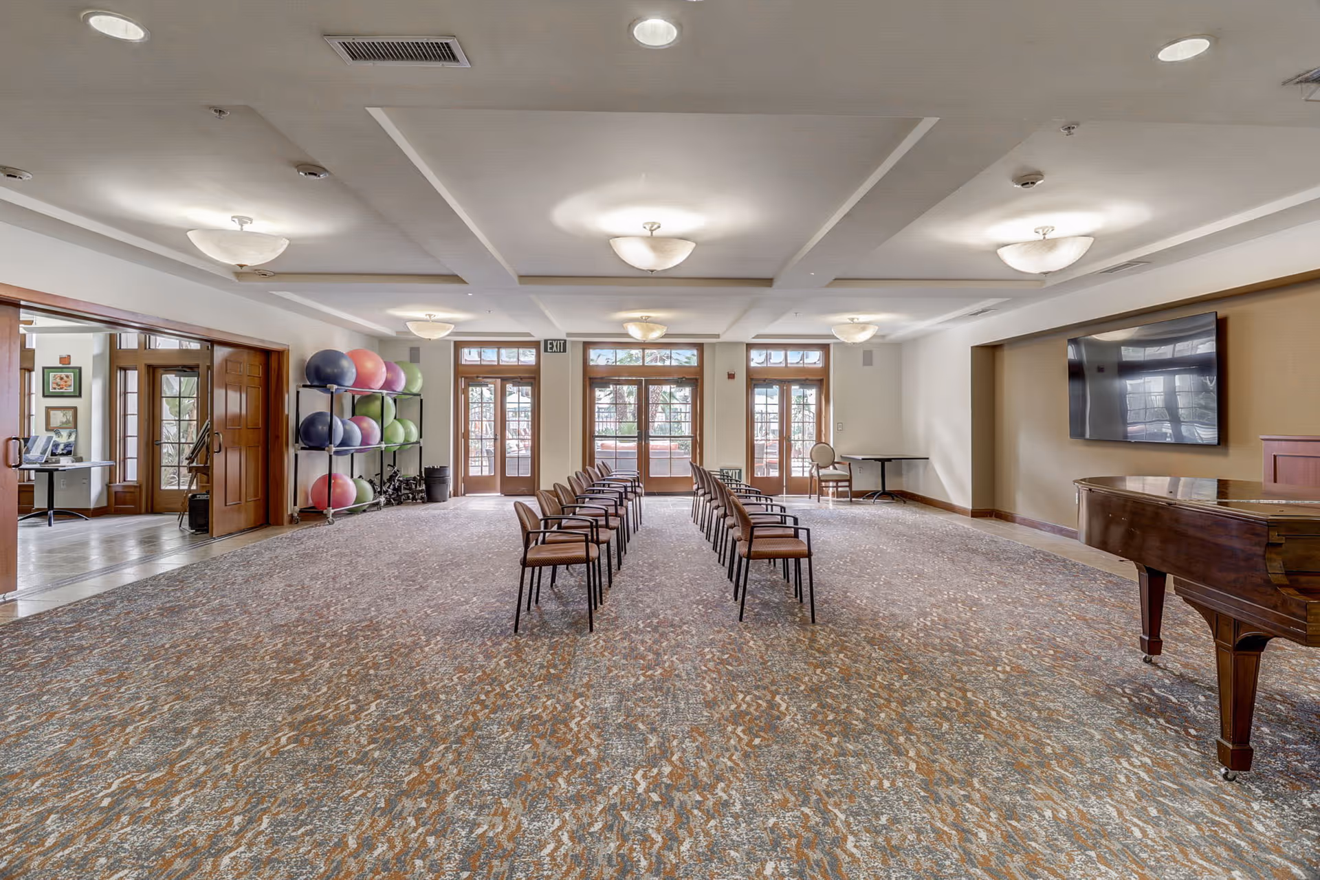 A spacious room with two rows of chairs facing a wall-mounted flat screen TV and a grand piano. The room has large windows and glass doors letting in natural light. On the left side, there is a rack holding colorful exercise balls. The ceiling has recessed lighting and decorative beams.