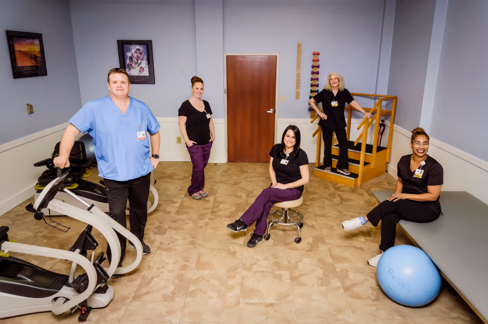 Five healthcare workers posing in a therapy or rehabilitation room. The room has exercise equipment, a therapy ball, and a small wooden staircase. The workers are wearing scrubs and name badges, smiling at the camera.