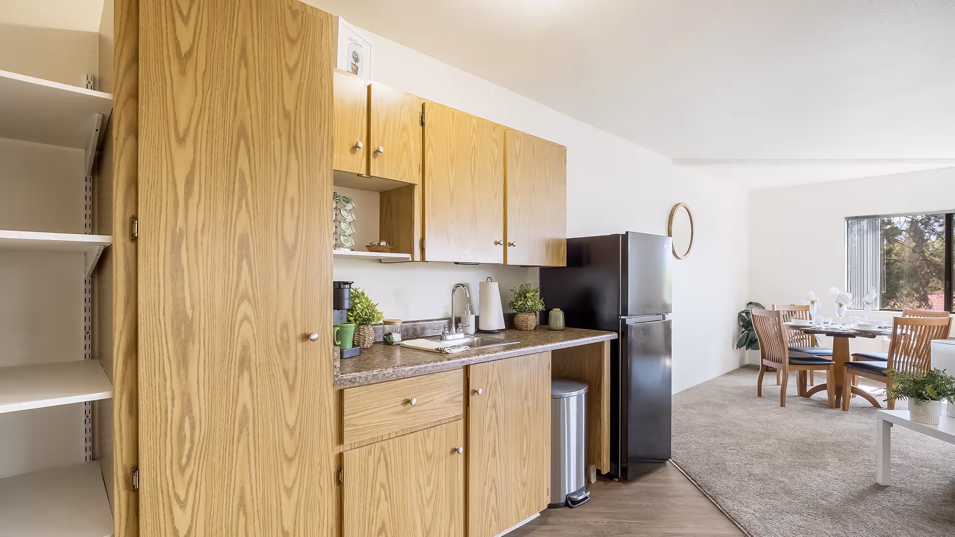 A bright and clean kitchen area with wooden cabinets, a countertop with a sink, a coffee maker, and small potted plants. To the right, there is a black refrigerator and a dining area with a round wooden table and four chairs near a large window letting in natural light.