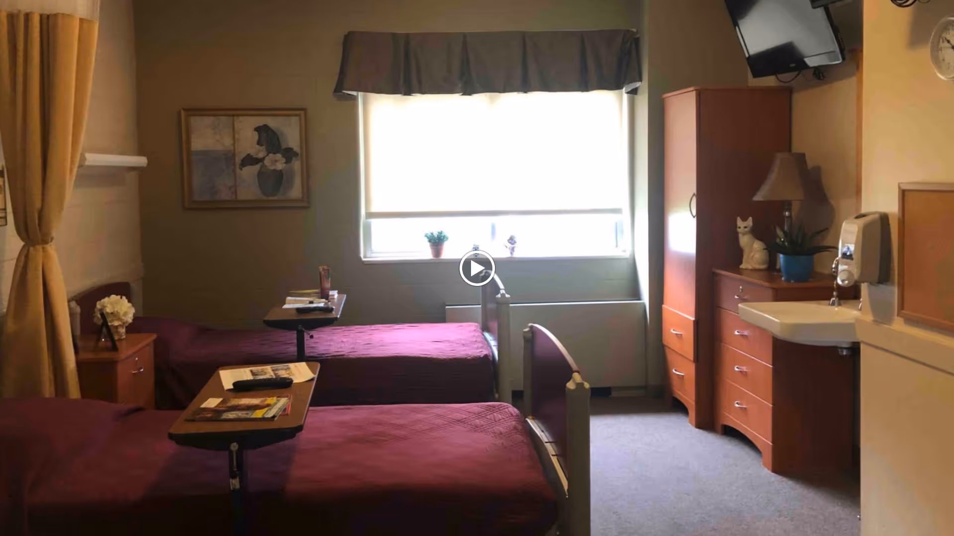 A senior living facility bedroom with two single beds covered in maroon bedspreads. Each bed has a small overbed table with items on top. The room has a window with a blind partially drawn, a wooden dresser with a lamp and decorative items, a wall-mounted TV, and a small sink with a soap dispenser.