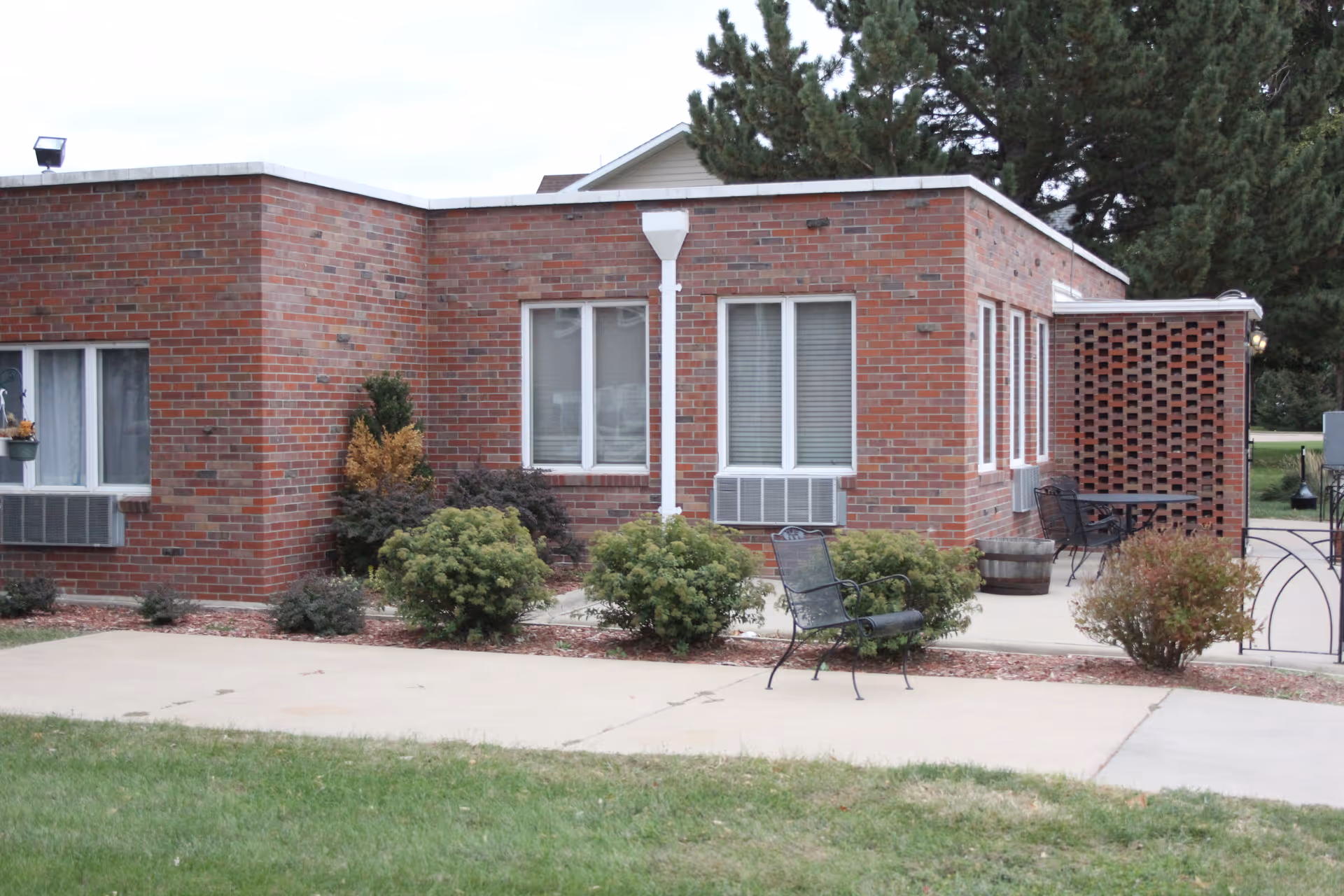 Single-story brick building facade with windows, shrubs, and metal patio chairs on a concrete walkway.