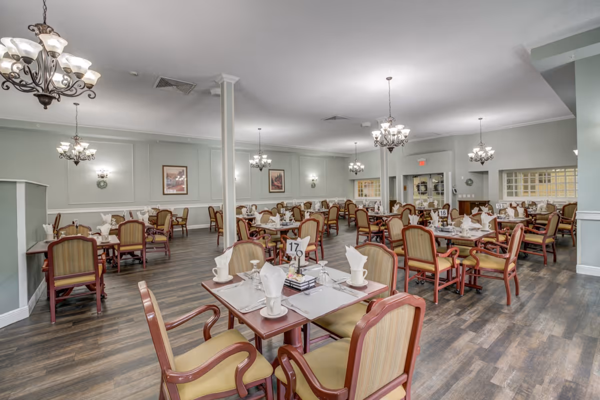 A spacious dining room with multiple tables set for meals, each table having four chairs with wooden frames and cushioned seats. The tables are arranged neatly with white napkins, cups, and glasses. The room features soft green walls, several chandeliers hanging from the ceiling, framed artwork on the walls, and wood flooring.
