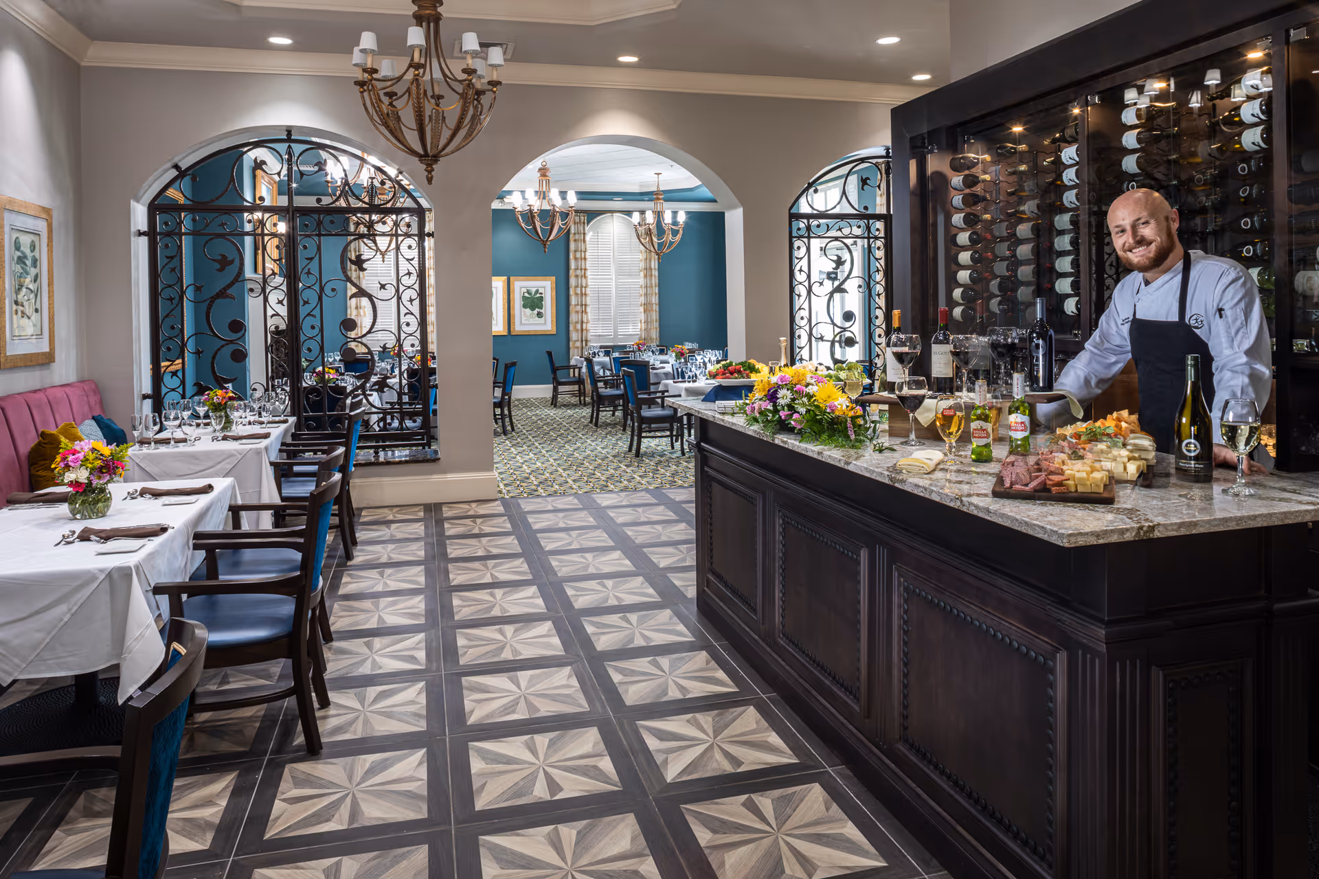A well-lit dining room with tables set with white tablecloths, glassware, and floral centerpieces. A smiling chef stands behind a marble countertop bar displaying various wines, beers, and a charcuterie board. The room features decorative wrought iron accents, chandeliers, and patterned flooring.