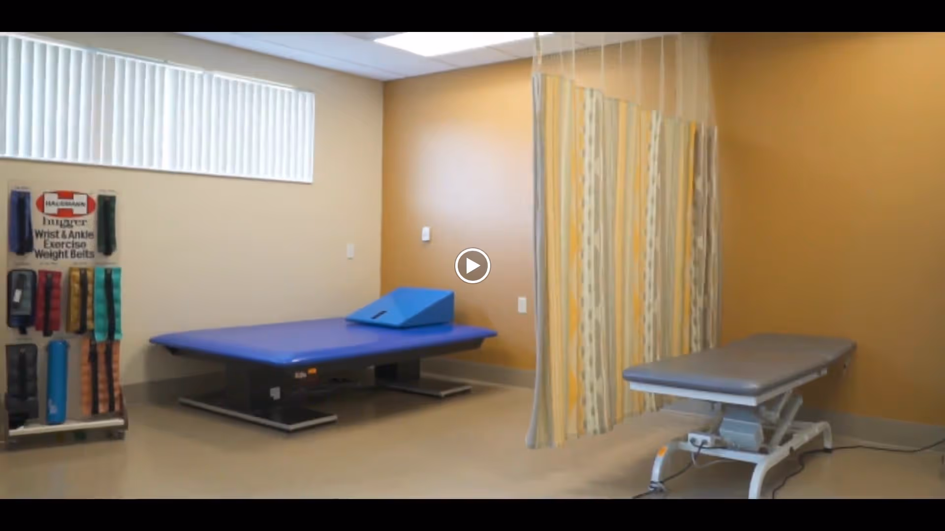 A therapy or treatment room with two adjustable treatment tables, one blue and one gray, separated by a yellow and gray patterned privacy curtain. On the left side, there is a rack holding various wrist and ankle exercise weight belts. The room has beige walls and a window with vertical blinds near the ceiling.