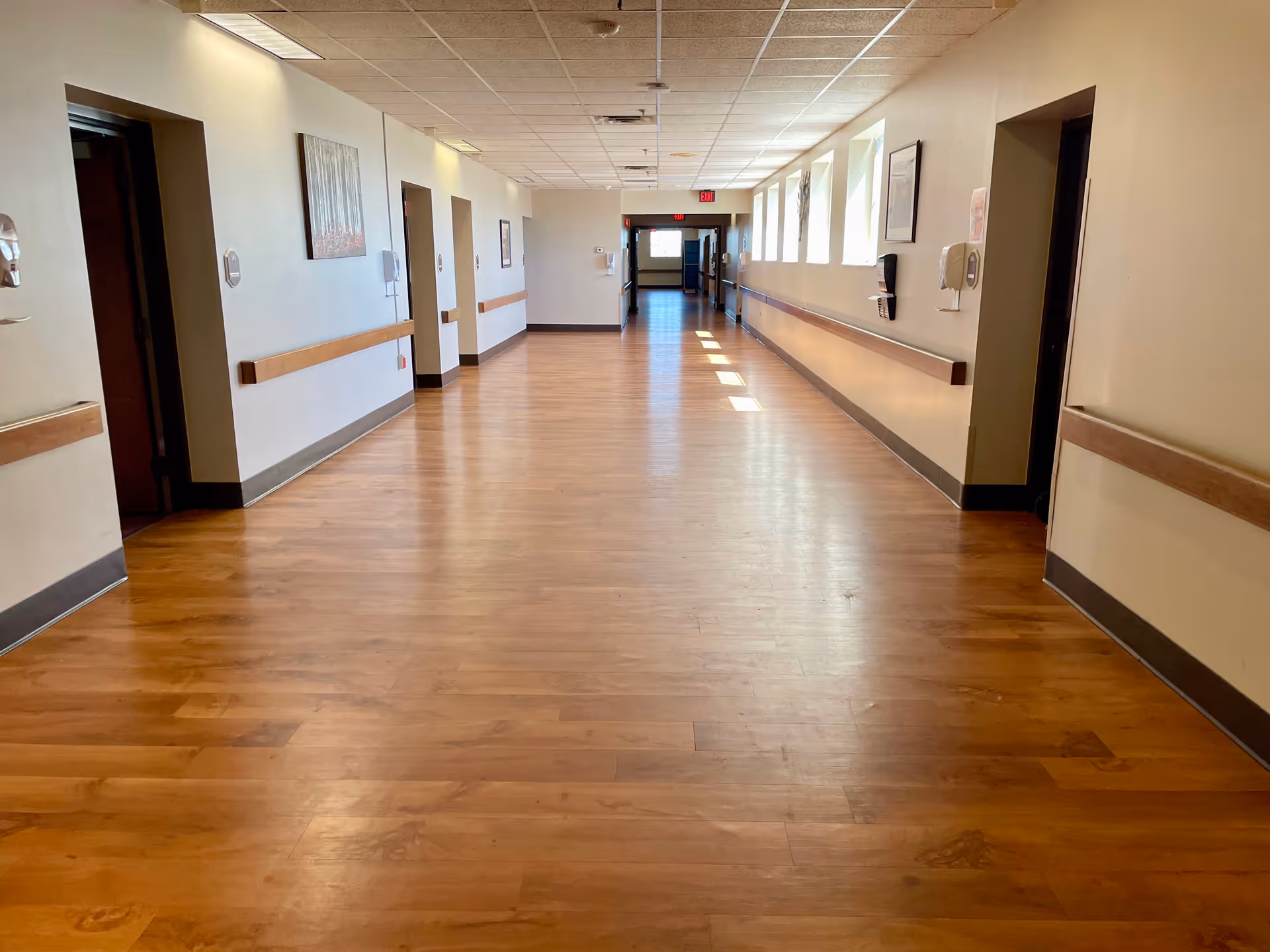 Long empty hallway in a care facility with wood-look floors, handrails along the walls, and doorways on both sides.