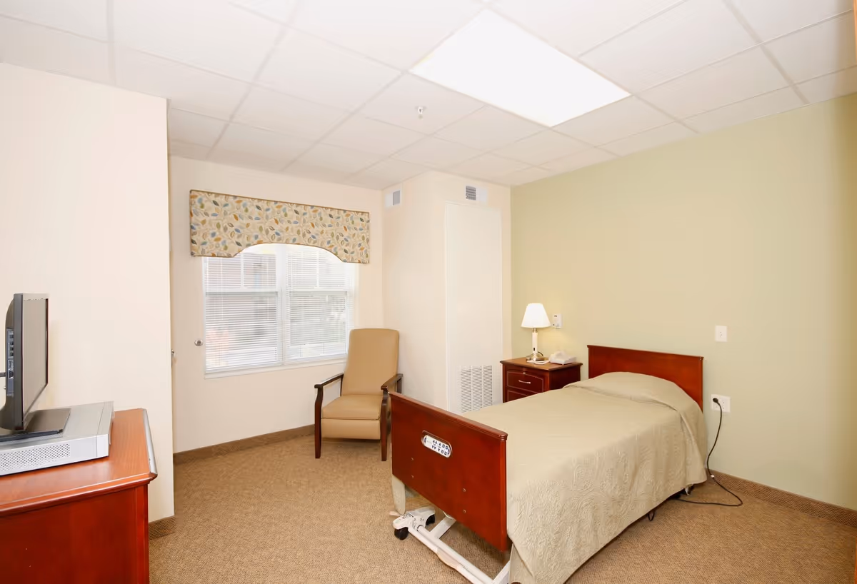 A simple senior living bedroom with a single bed covered in a beige quilt, a wooden nightstand with a lamp and telephone, a beige armchair near a window with patterned valance, and a TV on a wooden dresser.