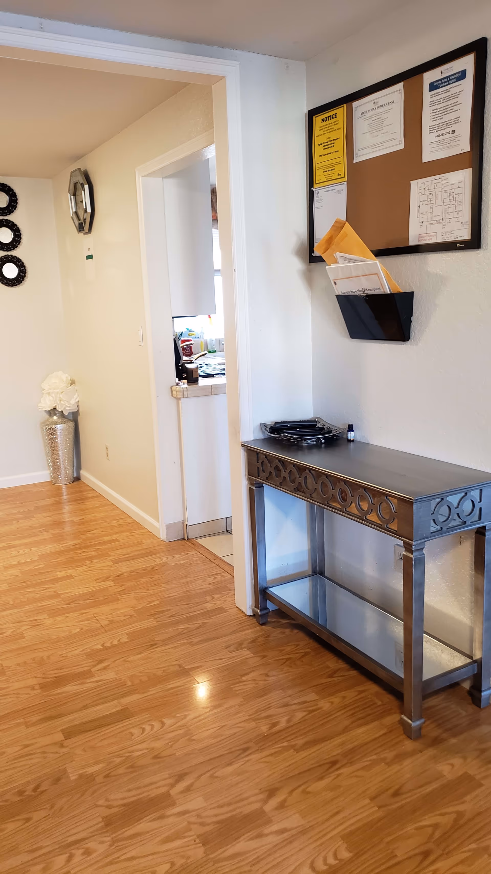 Interior hallway with wooden flooring, a decorative silver table with a glass shelf, a wall-mounted bulletin board with papers and notices, and a doorway leading to a kitchen area. There is also a silver vase with white flowers and decorative wall mirrors on the left wall.