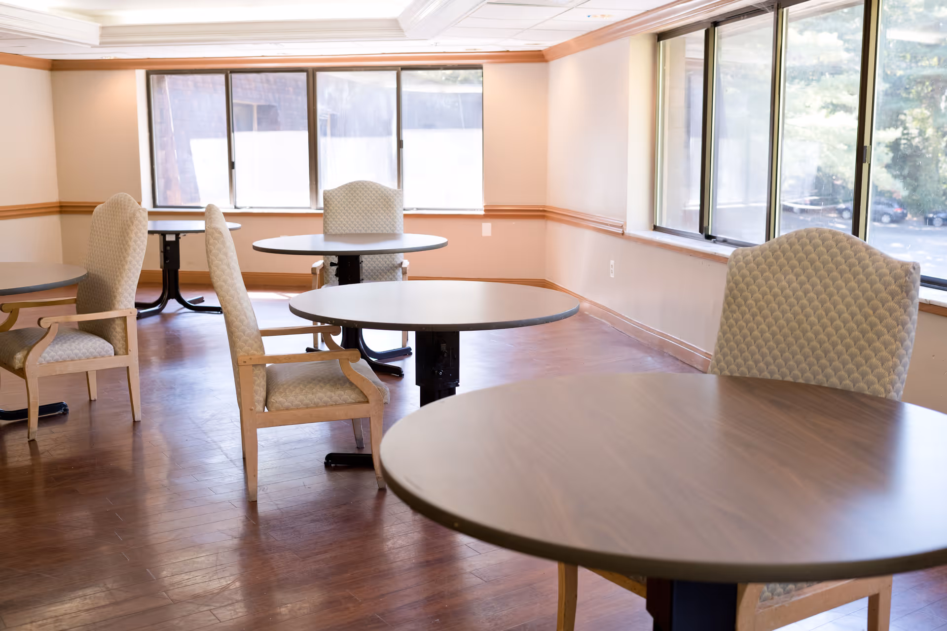 A bright dining area with round wooden tables and cushioned chairs with armrests. Large windows let in natural light and offer a view of trees and a parking lot outside. The room has wooden flooring and beige walls with wooden trim.