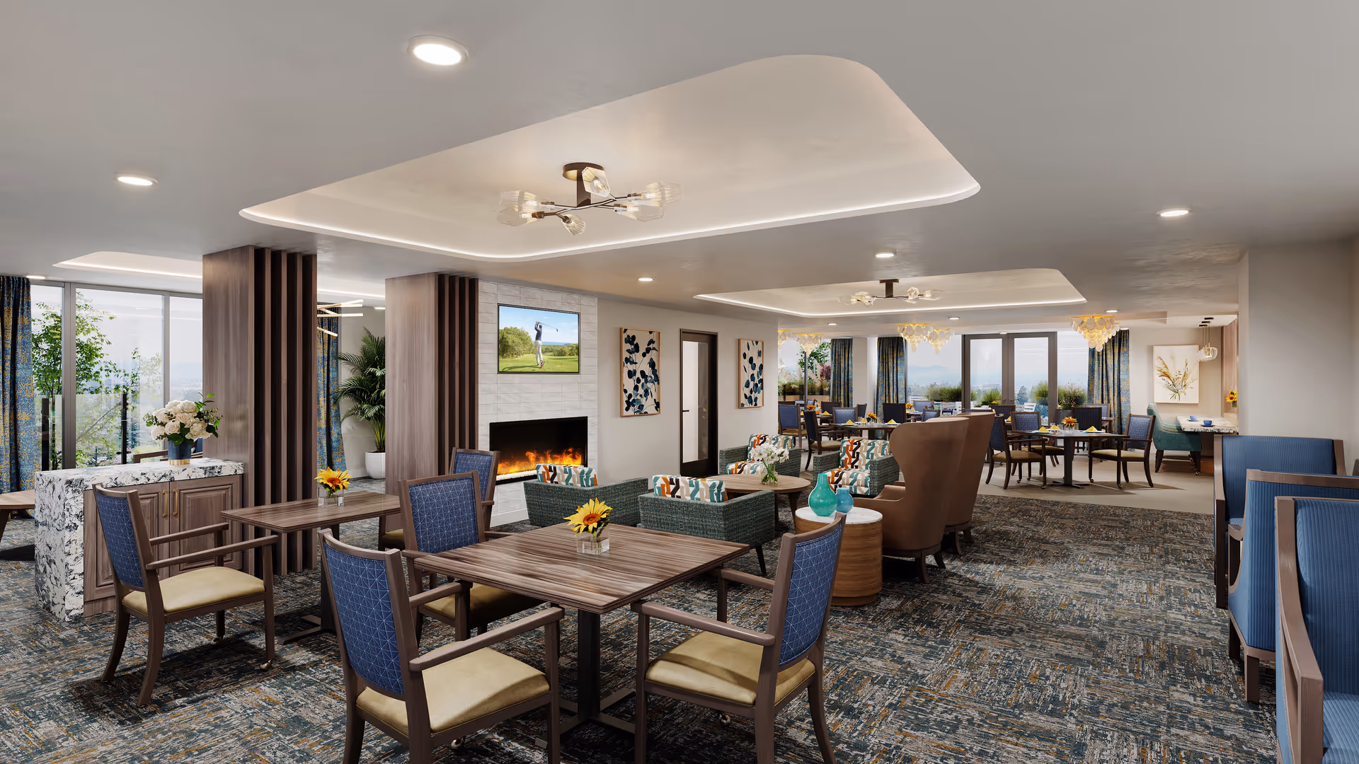 A spacious and well-lit common area in a senior living facility featuring multiple seating arrangements with wooden tables and cushioned chairs. The room includes a cozy fireplace with a TV mounted above it, decorative wall art, and large windows allowing natural light to fill the space. The area is carpeted with a patterned design and has modern ceiling lights and chandeliers.