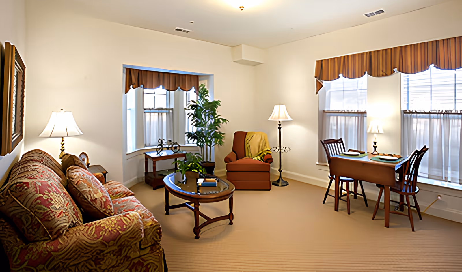 A cozy living room with a patterned sofa, a glass-top coffee table, a potted plant, an armchair with a throw blanket, and a small dining table set for two near windows with brown valances and sheer curtains. The room is softly lit by multiple lamps.