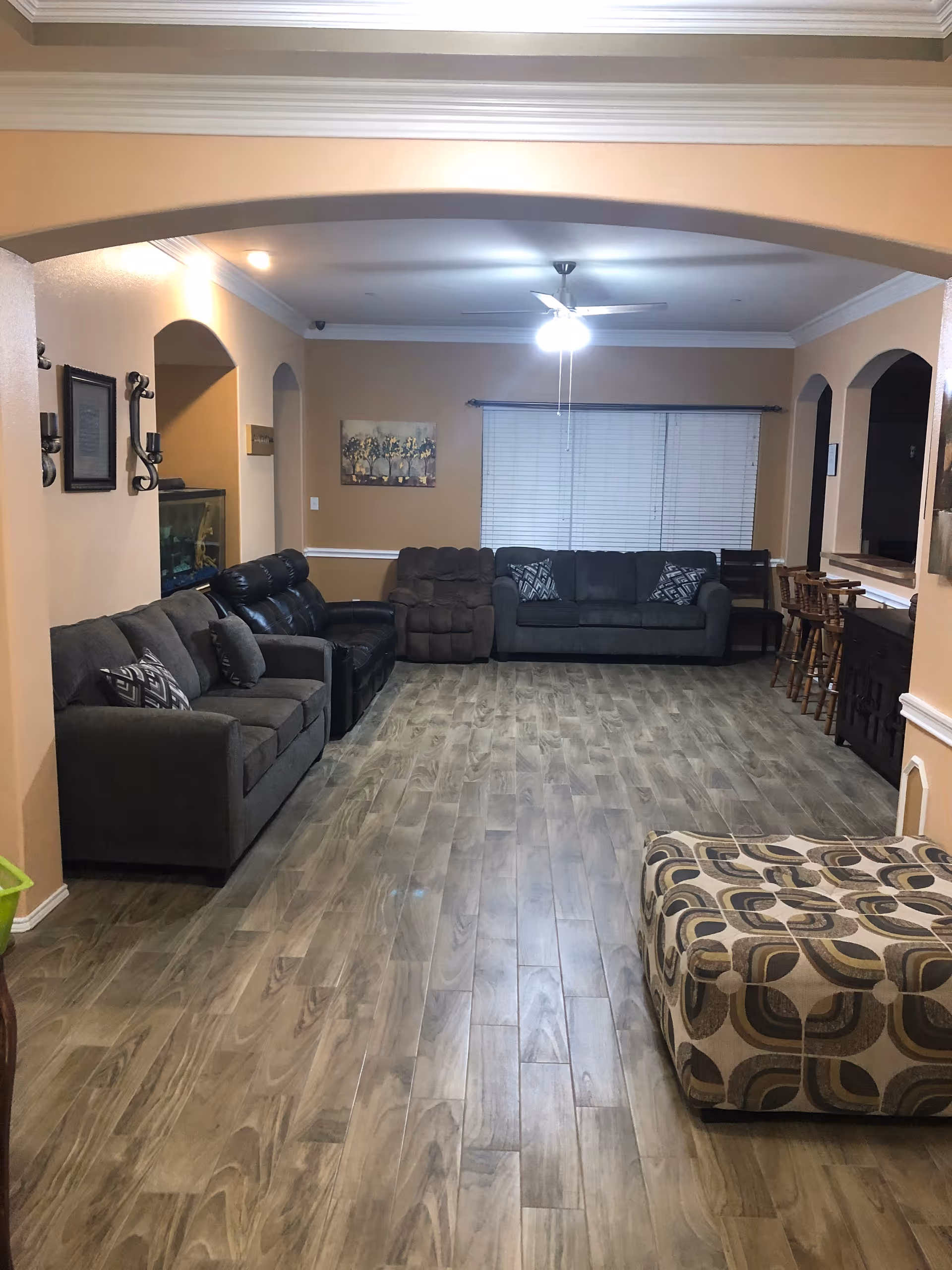 A spacious living room with wooden flooring, beige walls, and white crown molding. The room features multiple seating options including a gray sofa, a black leather recliner, a dark gray sofa, and a patterned ottoman. There is a ceiling fan with lights in the center of the room, a window with closed blinds on the far wall, and a row of bar stools along a counter on the right side. Wall decorations include framed pictures and sconces.