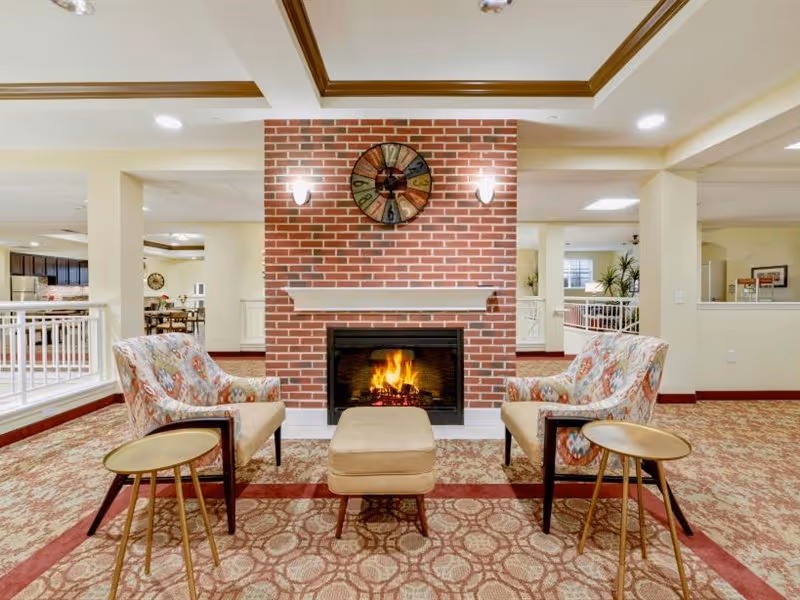 A cozy seating area in a senior living facility featuring a brick fireplace with a fire burning inside. Two patterned armchairs face each other with a beige ottoman in between. Each chair has a small round side table next to it. Above the fireplace is a colorful wall clock. The room has patterned carpet, cream-colored walls, and recessed lighting in the ceiling.