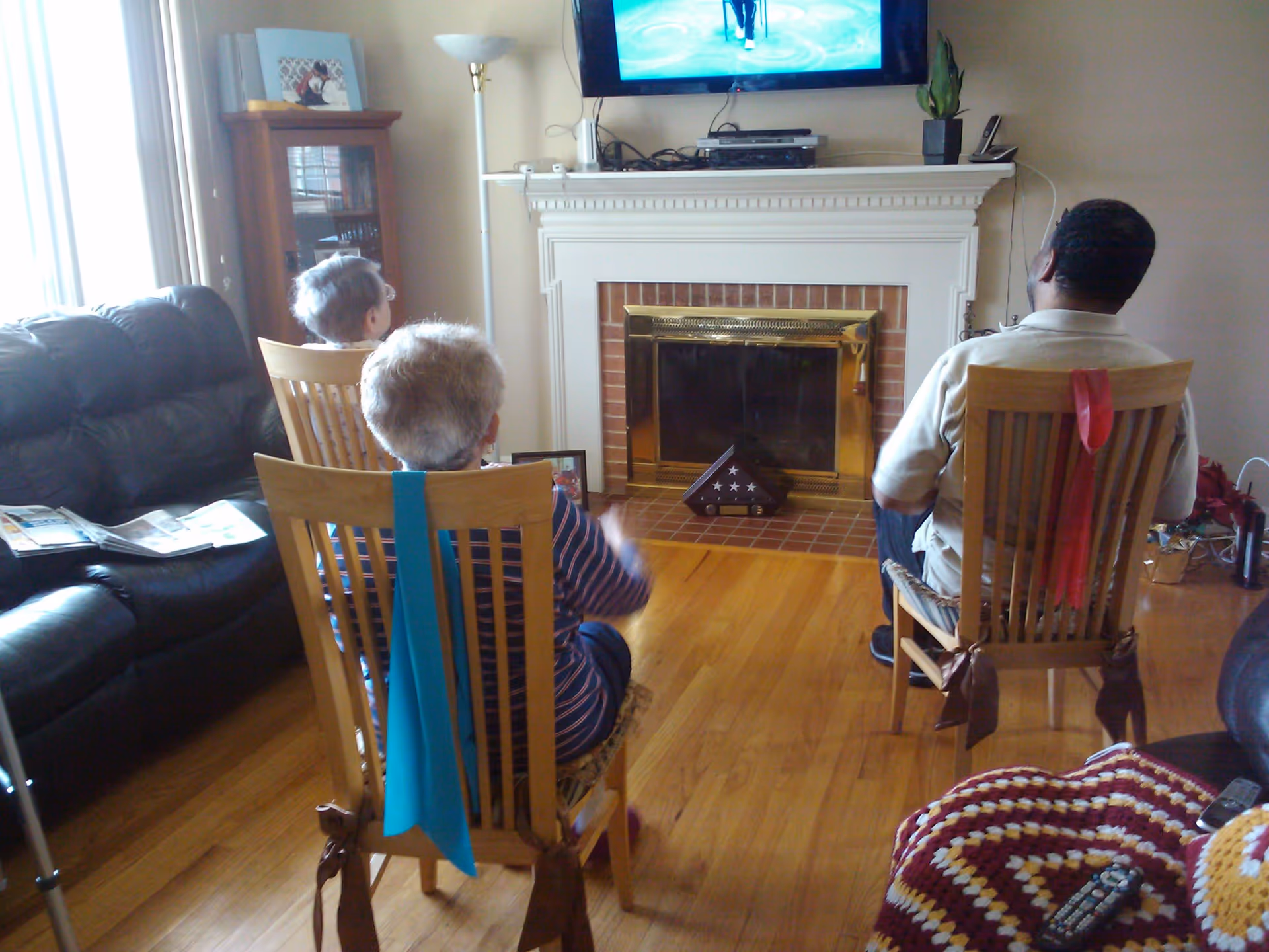 Three elderly people sitting on wooden chairs in a living room, facing a television mounted above a white fireplace with a brass screen. The room has hardwood floors, a black leather couch on the left, a wooden cabinet with framed photos, and a crocheted blanket on a piece of furniture in the foreground. Two of the chairs have colored fabric strips tied to the backrests.