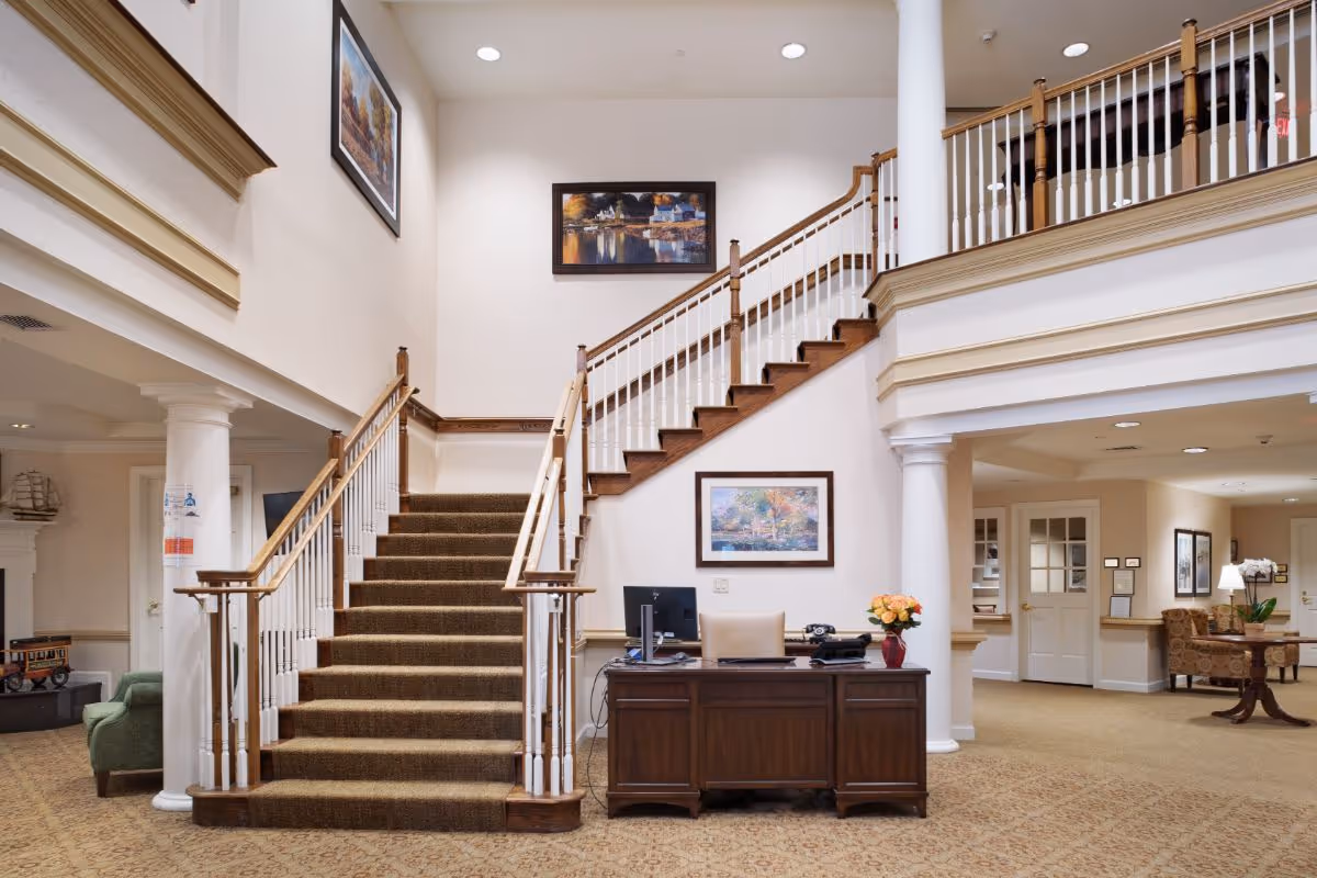 Interior view of a senior living facility lobby featuring a carpeted staircase with wooden handrails leading to an upper floor. A wooden reception desk with a computer, phone, and a vase of flowers is positioned in front of the staircase. The walls are decorated with framed paintings, and there are white columns and beige carpeting throughout the space. Seating areas with chairs and tables are visible in the background.