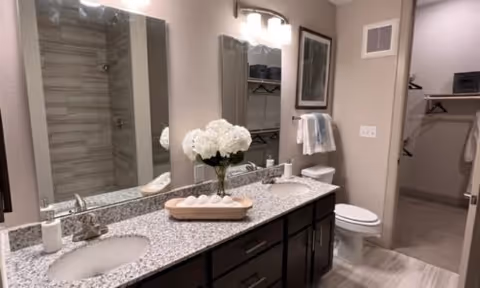 Modern bathroom with a granite countertop featuring two sinks, a large mirror, a vase with white flowers, and a tray with rolled white towels. The bathroom has a toilet, a towel rack with a folded towel, and an open doorway leading to a walk-in closet.