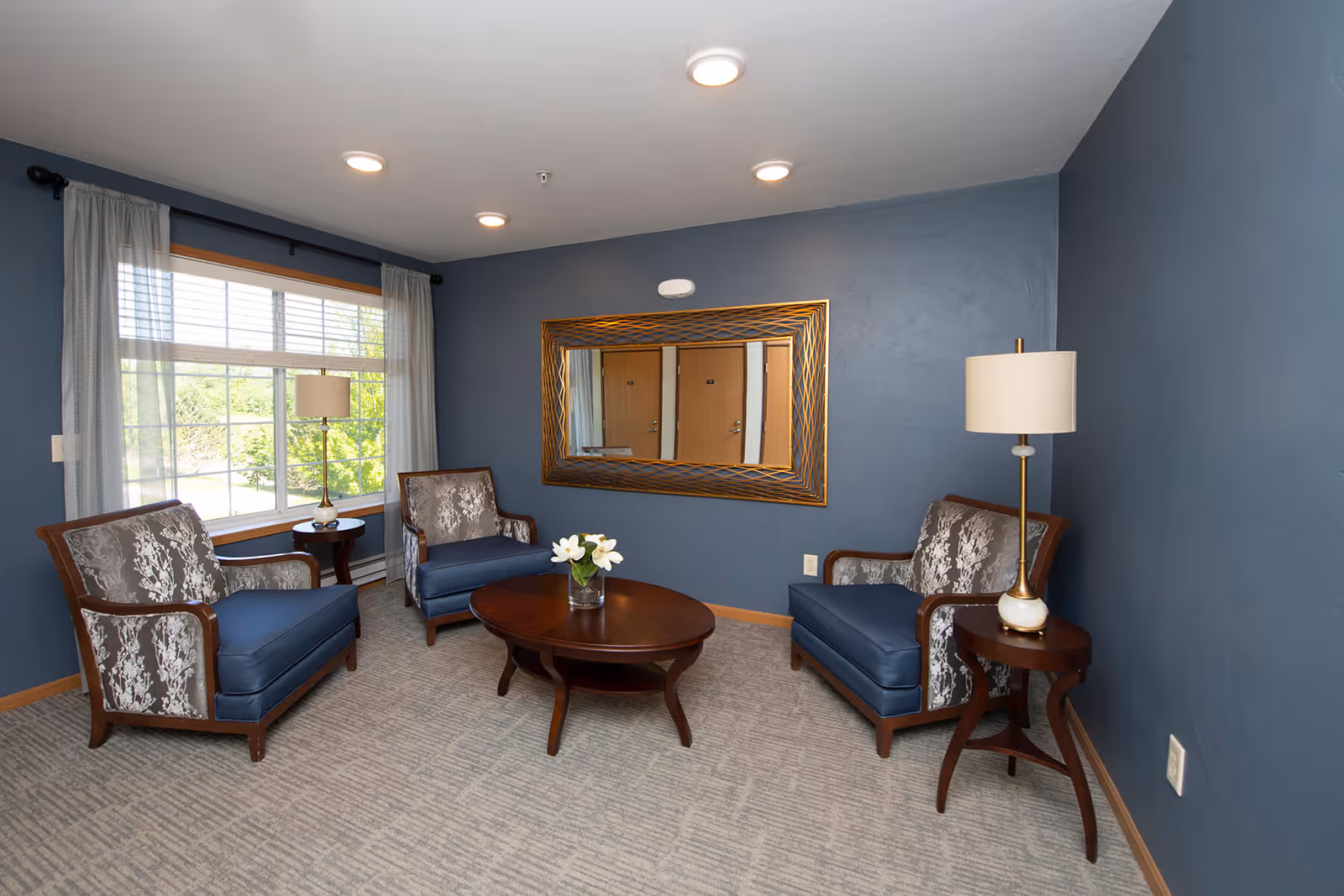 Small sitting room with three upholstered armchairs around a round coffee table, a large window, wall mirror, and floor lamps.