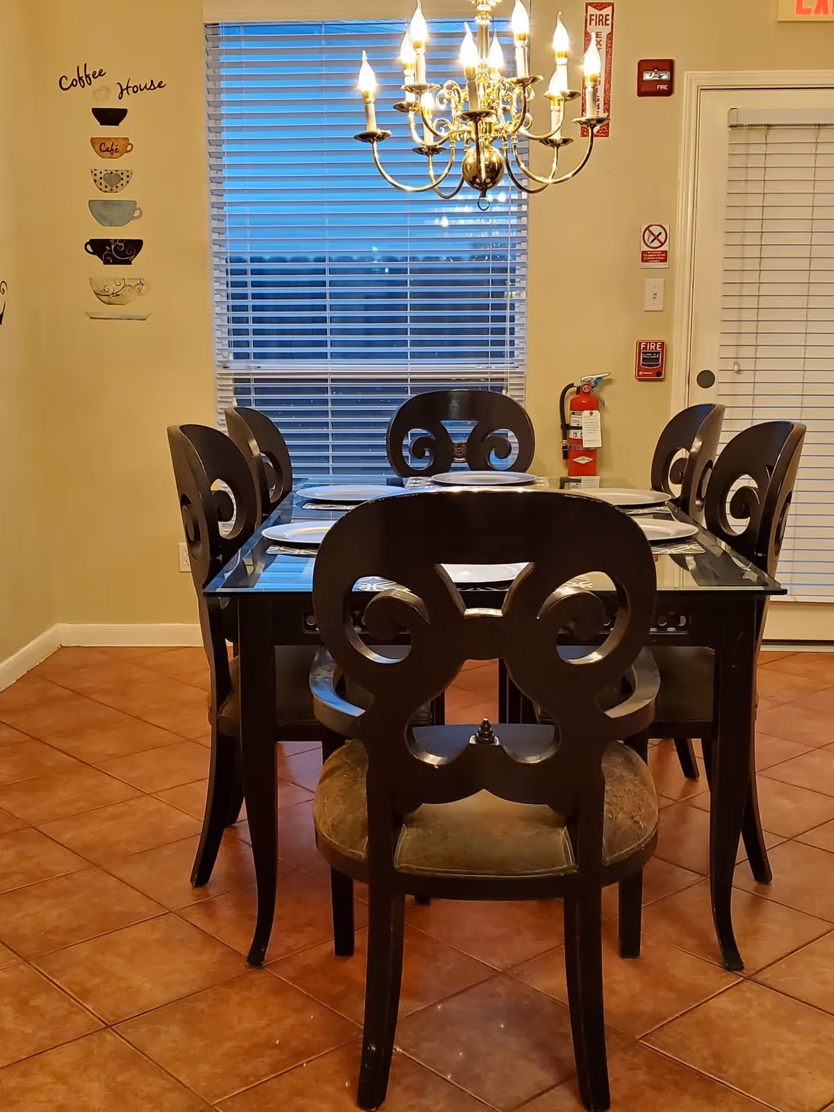 A dining room with a glass-top table and six decorative chairs set with plates beneath a chandelier, with window blinds and a tiled floor.