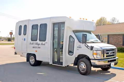 White shuttle bus parked outside a brick senior living facility labeled North Village Park.