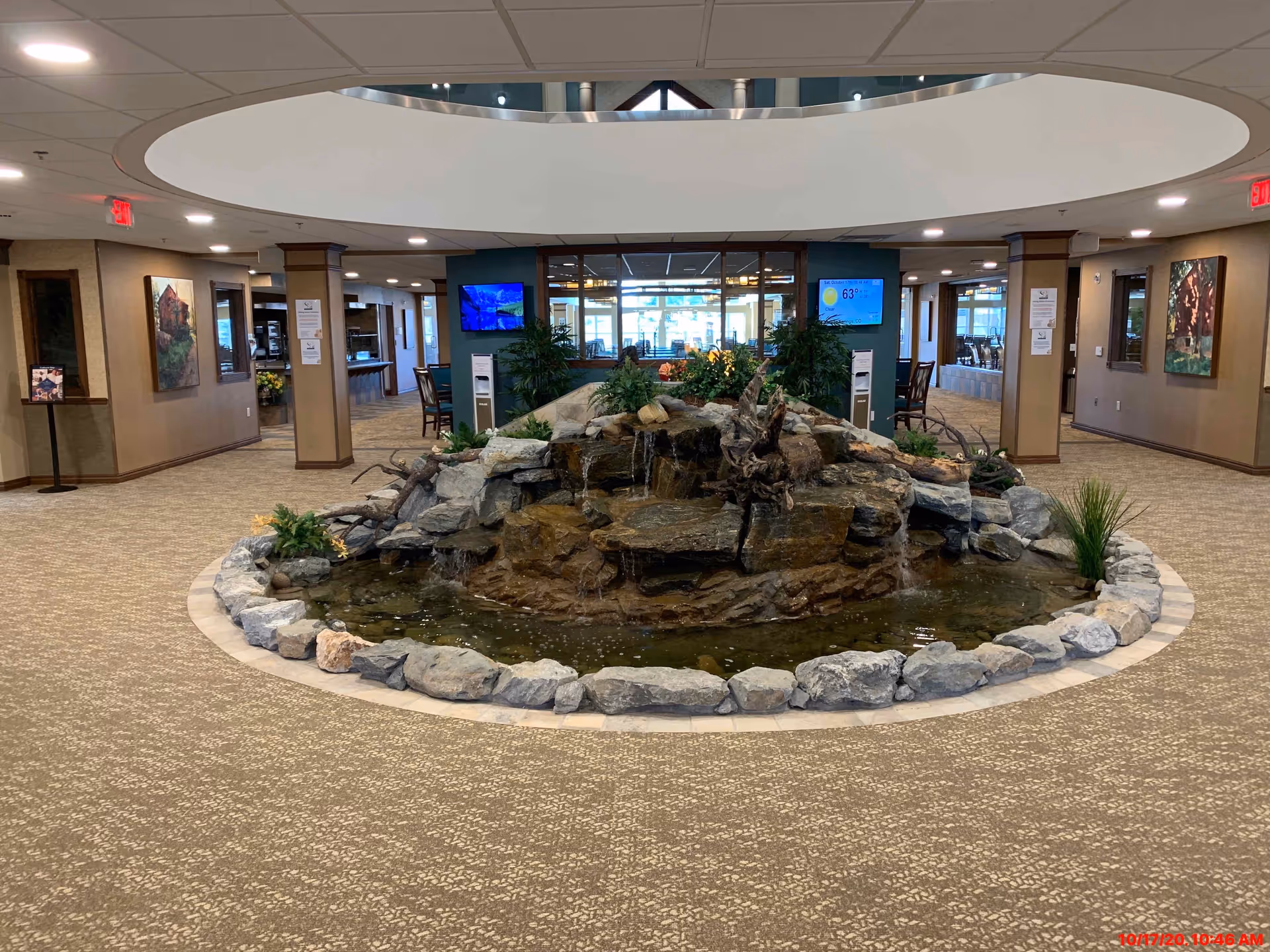 Indoor area of a senior living facility featuring a large circular rock water fountain in the center surrounded by carpeted flooring. The space has beige walls with framed artwork, several pillars, and seating areas visible in the background. Two digital screens display weather and other information on the far wall.