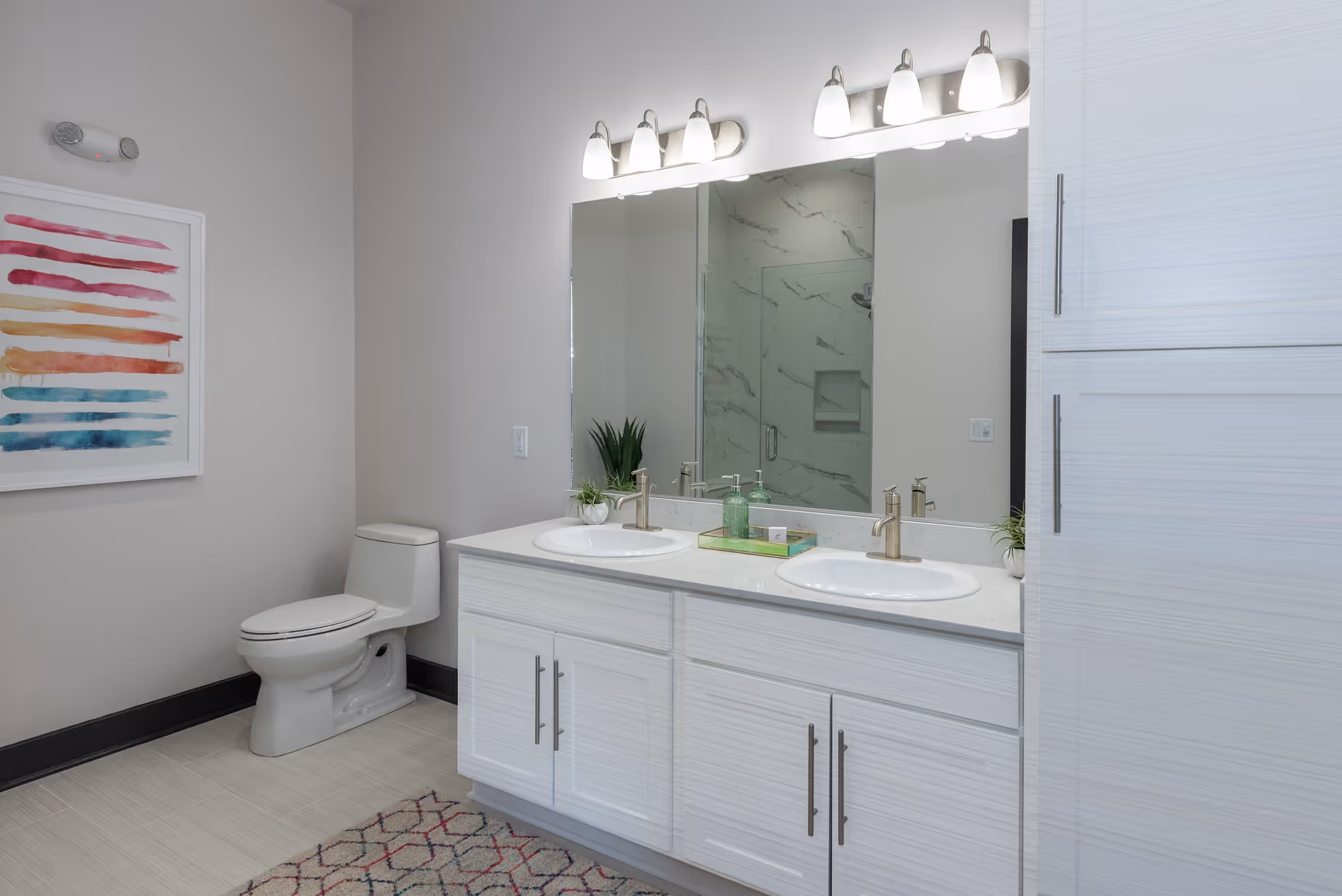 A modern bathroom featuring a white toilet, a double sink vanity with white cabinets and silver handles, a large mirror above the sinks with two sets of three-light fixtures, a glass-enclosed shower with marble-patterned tiles reflected in the mirror, a colorful abstract painting on the wall, and a patterned rug on the tiled floor.
