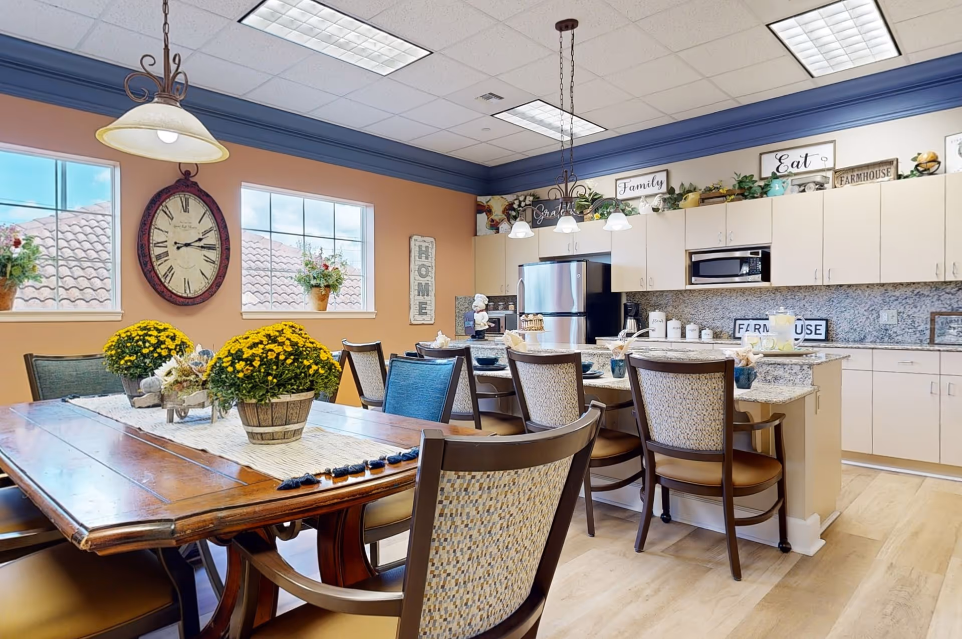 A bright and spacious kitchen and dining area with a wooden dining table decorated with yellow flowers in pots. The kitchen features light-colored cabinets, granite countertops, stainless steel refrigerator, microwave, and a kitchen island with seating. The walls are painted a warm peach color with two windows showing a tiled roof outside. Decorative signs with words like 'HOME', 'Family', 'Eat', and 'Farmhouse' are displayed above the cabinets.