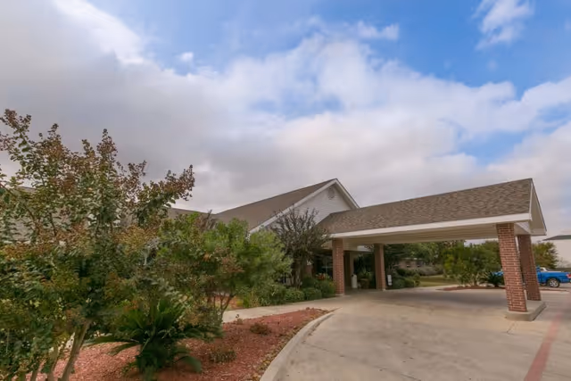 Exterior view of Legend Oaks Healthcare and Rehabilitation - South San Antonio showing the building entrance with a covered driveway, surrounded by landscaped greenery and trees under a partly cloudy sky.