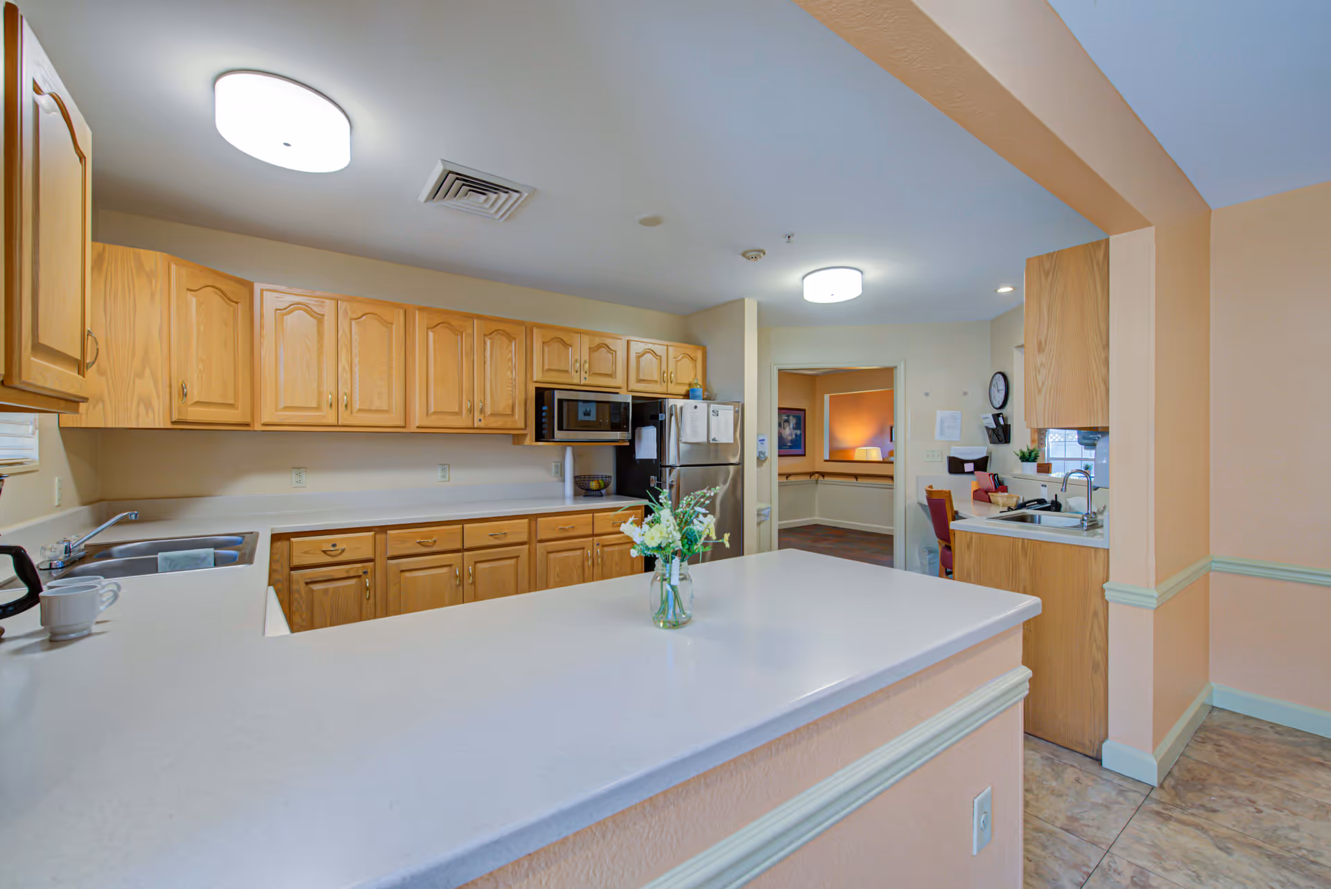 A clean and well-lit kitchen area with light wood cabinets, a white countertop island with a small vase of flowers, a stainless steel refrigerator, microwave, and a double sink. The walls are painted a soft peach color, and there is a doorway leading to another room with a chair and wall decorations visible.