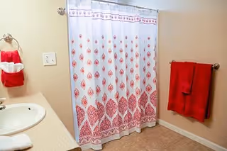 A bathroom with a white sink and countertop on the left, a red and white patterned shower curtain in the center, and red towels hanging on the wall to the right and near the sink. The walls are beige and the floor is tiled.