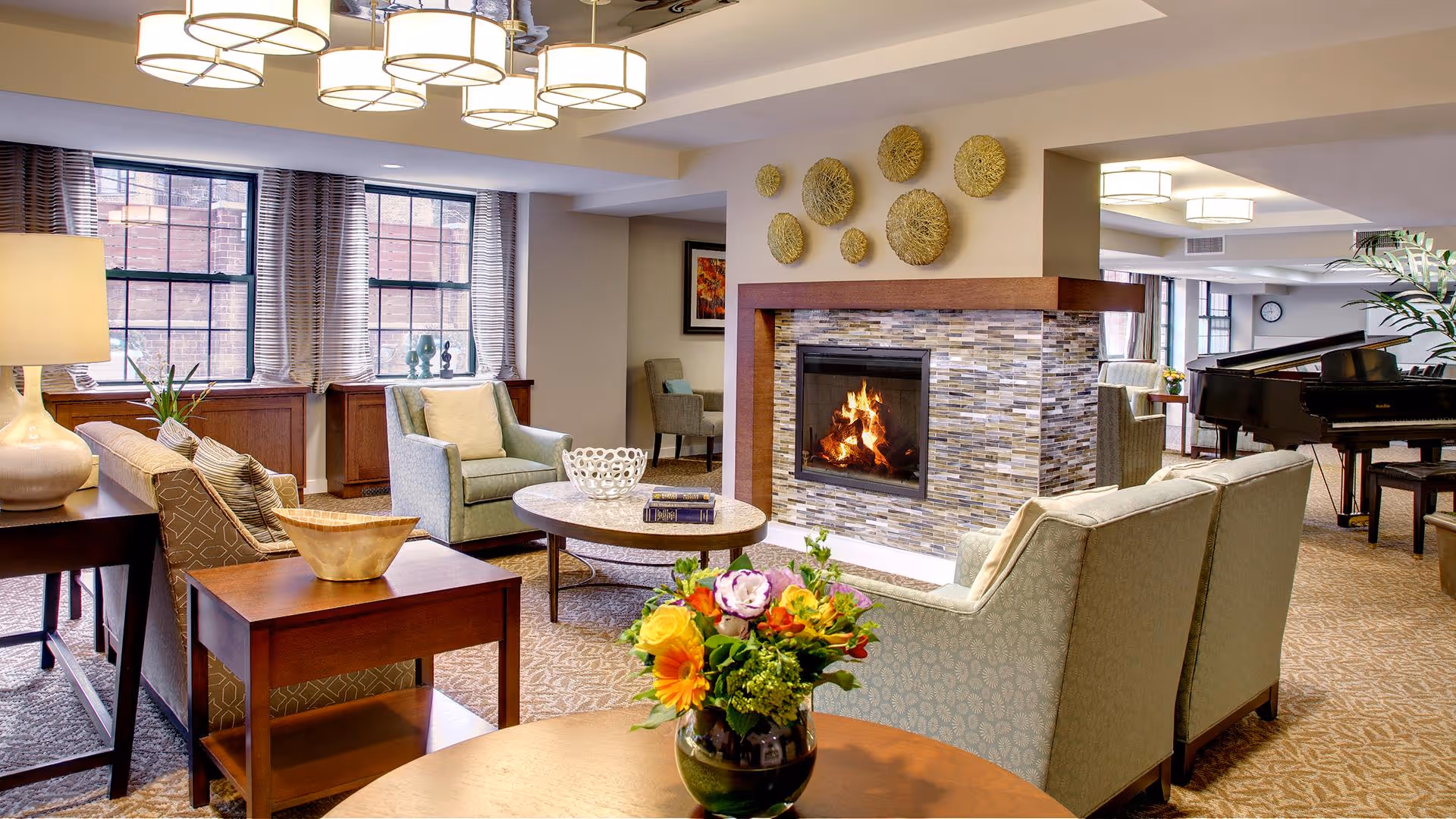 A cozy living room area in a senior living facility featuring a lit fireplace with decorative wall art above it, comfortable armchairs and sofas arranged around a round coffee table, a side table with a lamp and decorative bowl, large windows with curtains, and a grand piano in the background. A vase with colorful flowers is placed on a round table in the foreground.