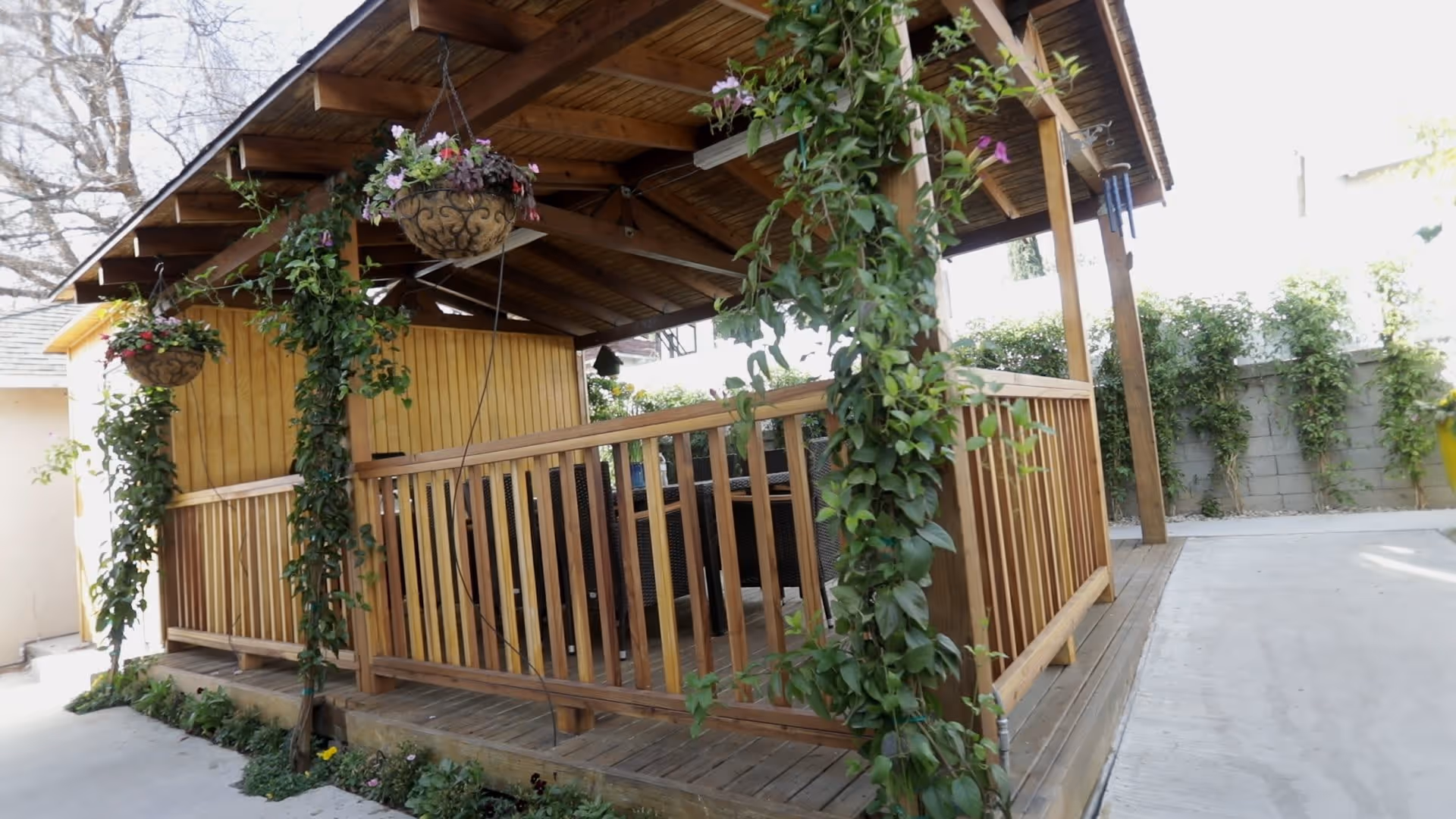 Outdoor wooden gazebo structure with a slanted roof, surrounded by hanging flower pots and climbing plants on the wooden posts. The area around the gazebo is paved with concrete and there are green shrubs along a concrete wall in the background.