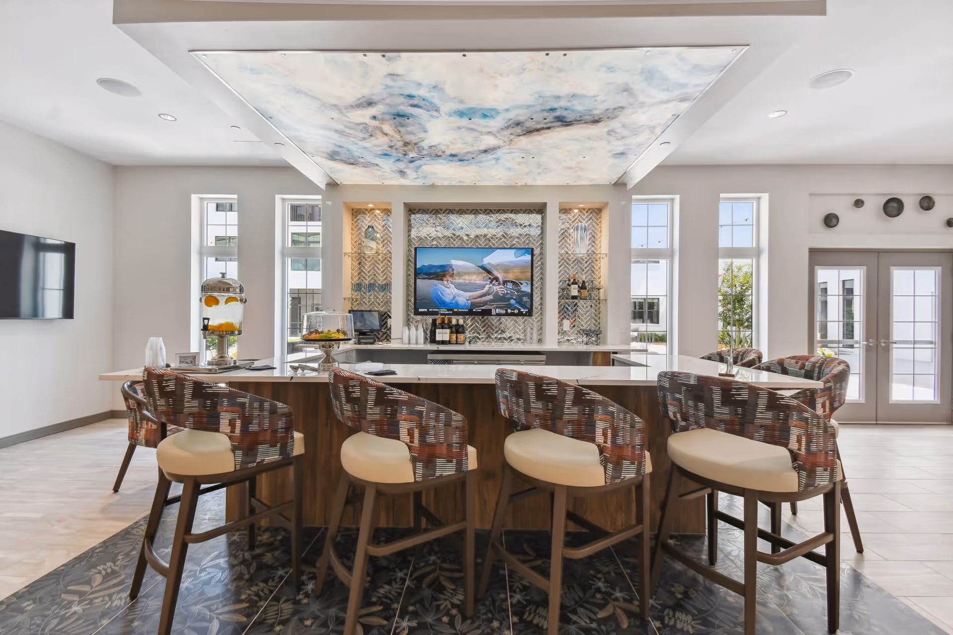 Modern senior living facility common area with a large island bar surrounded by six patterned cushioned bar stools. The bar area features a mounted flat-screen TV, decorative shelving with bottles and glassware, and a ceiling panel with a cloud-like design. Large windows and glass doors allow natural light to fill the space.