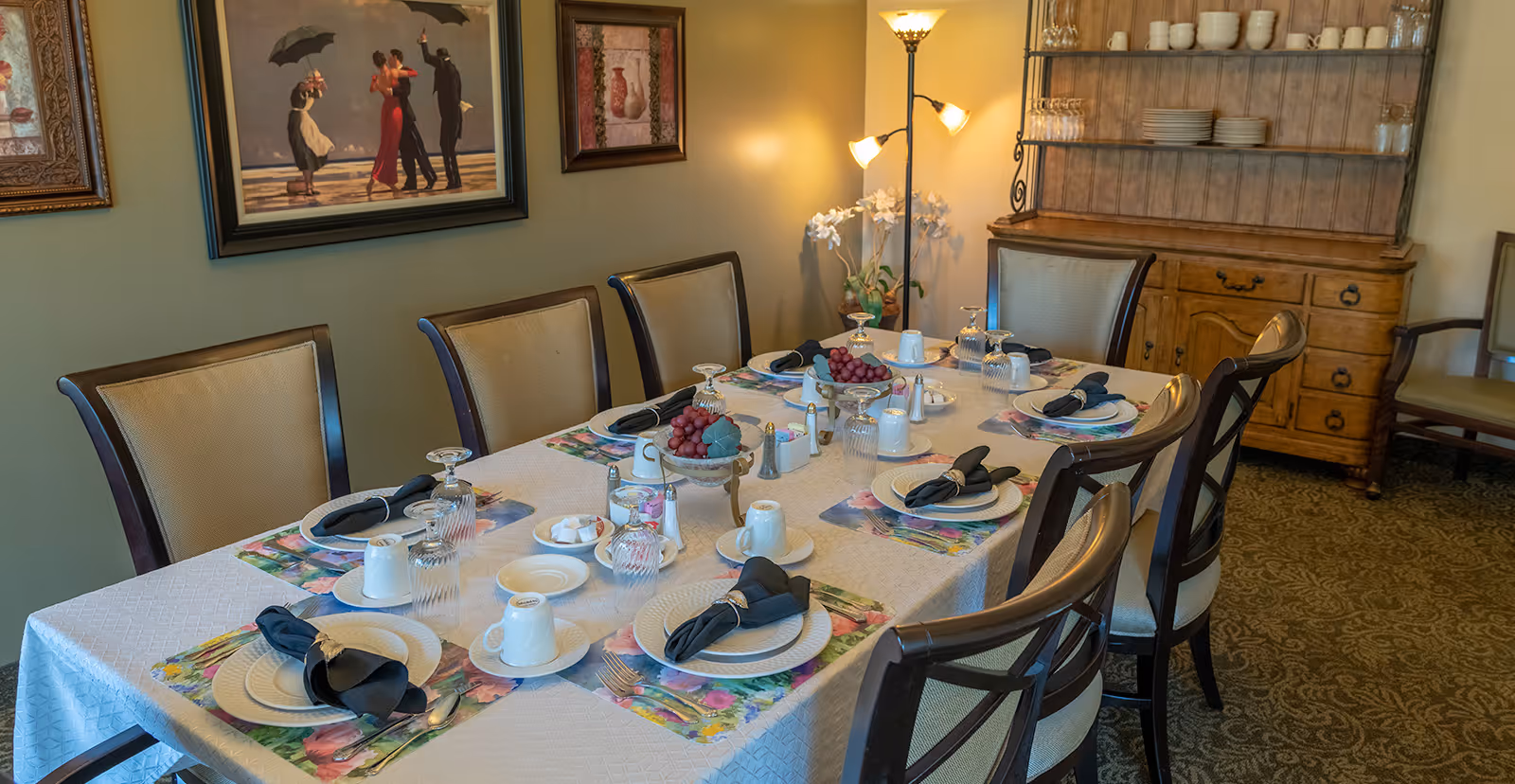 A dining room with a rectangular table set for eight people. The table is covered with a white tablecloth and colorful placemats, each setting includes a plate, a black cloth napkin with a napkin ring, a cup on a saucer, and an upside-down glass. There are two bowls of red grapes in the center of the table along with salt and pepper shakers and sugar packets. The room has beige walls with framed artwork, a wooden cabinet with dishes and glasses, a floor lamp, and upholstered chairs around the table.