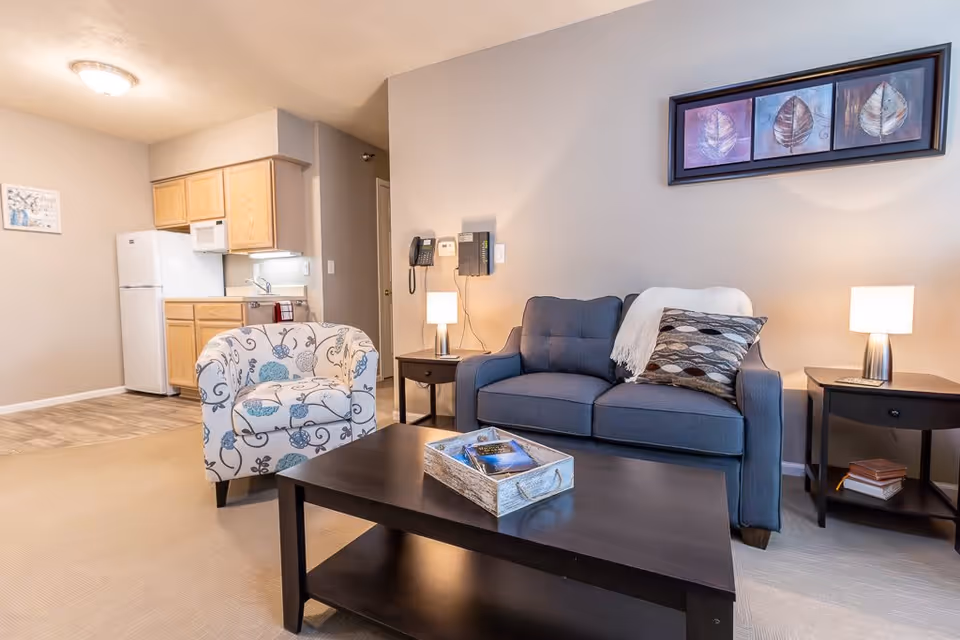 A cozy living room area with a blue upholstered sofa adorned with a patterned pillow and a white throw blanket. Next to the sofa are two dark wooden side tables, each with a small lamp. A patterned armchair with blue and gray floral designs is positioned near a dark wooden coffee table that holds a decorative tray with books. In the background, there is a small kitchen area with light wood cabinets, a white refrigerator, and a microwave. The walls are painted beige, and a framed artwork featuring three leaves hangs above the sofa.