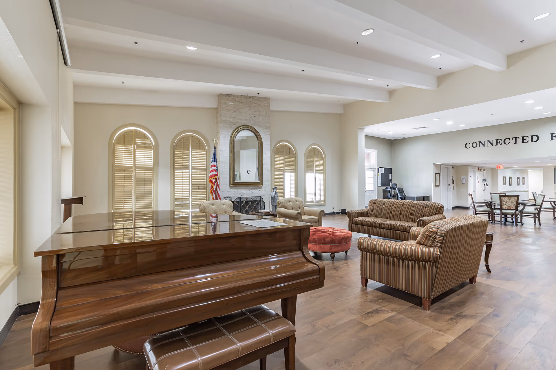 A spacious living area in a senior living facility featuring a polished wooden grand piano with a matching bench in the foreground. The room has a seating arrangement with striped sofas, a red round ottoman, and two beige armchairs near a fireplace with a large mirror above it. Three tall arched windows with shutters and an American flag are visible behind the fireplace. In the background, there is an open dining area with tables and chairs, and a wall with the words 'CONNECTED F' partially visible.