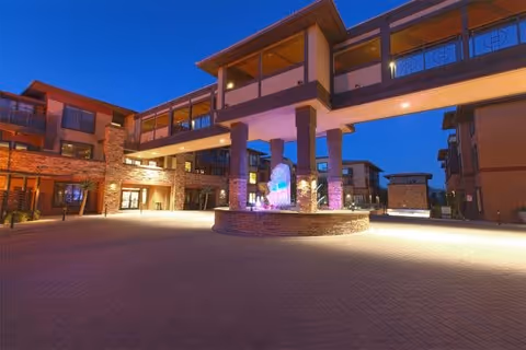 Exterior view of Andara By Cogir facility at dusk, showing a spacious courtyard with a central illuminated water fountain, surrounded by multi-story buildings connected by an elevated walkway.