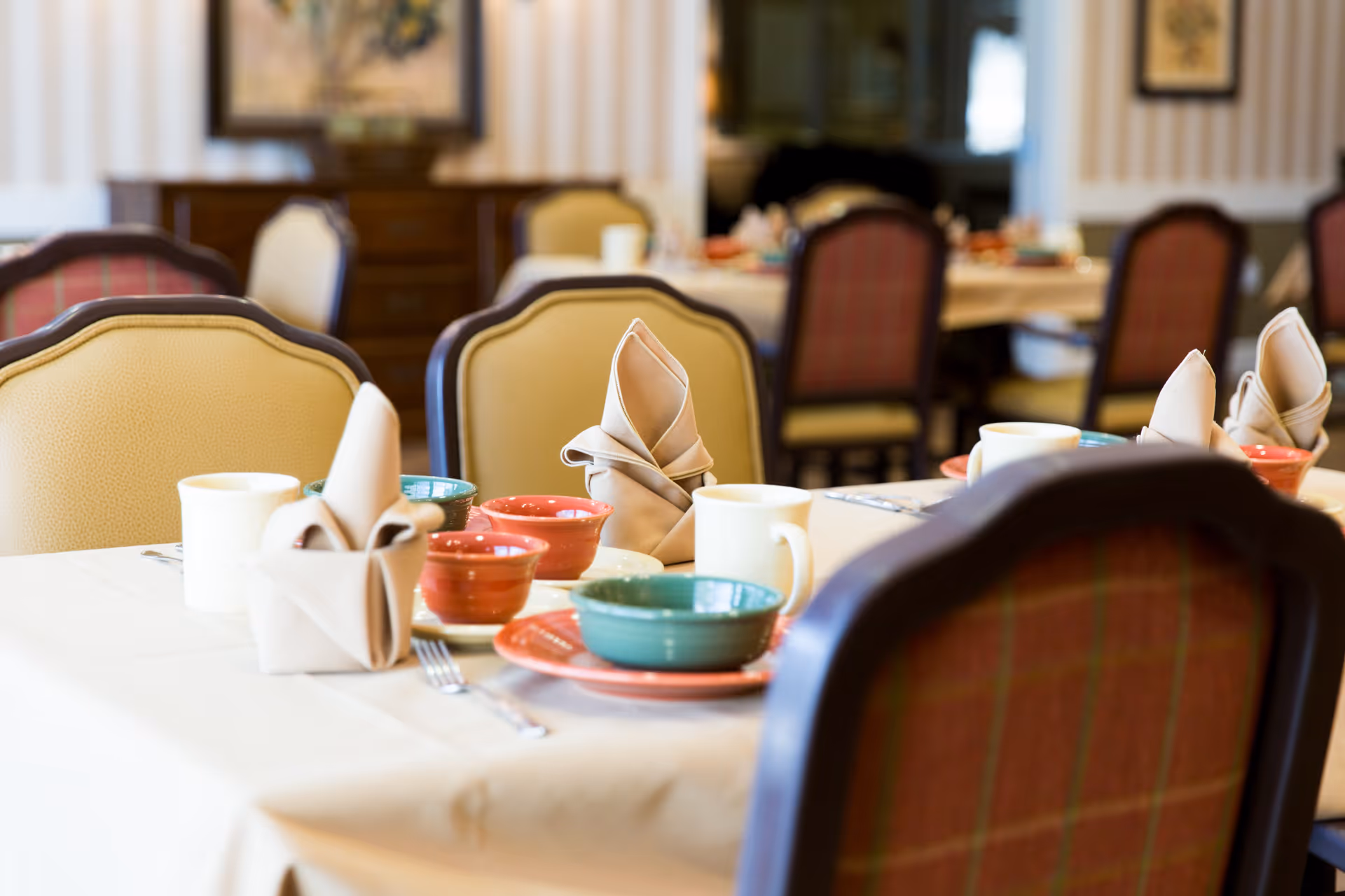 A dining room table set with folded beige napkins, colorful bowls, plates, and white mugs. The chairs around the table have cushioned backs with a mix of beige and red plaid upholstery. The background shows more tables and chairs in a softly lit room with framed artwork on the walls.