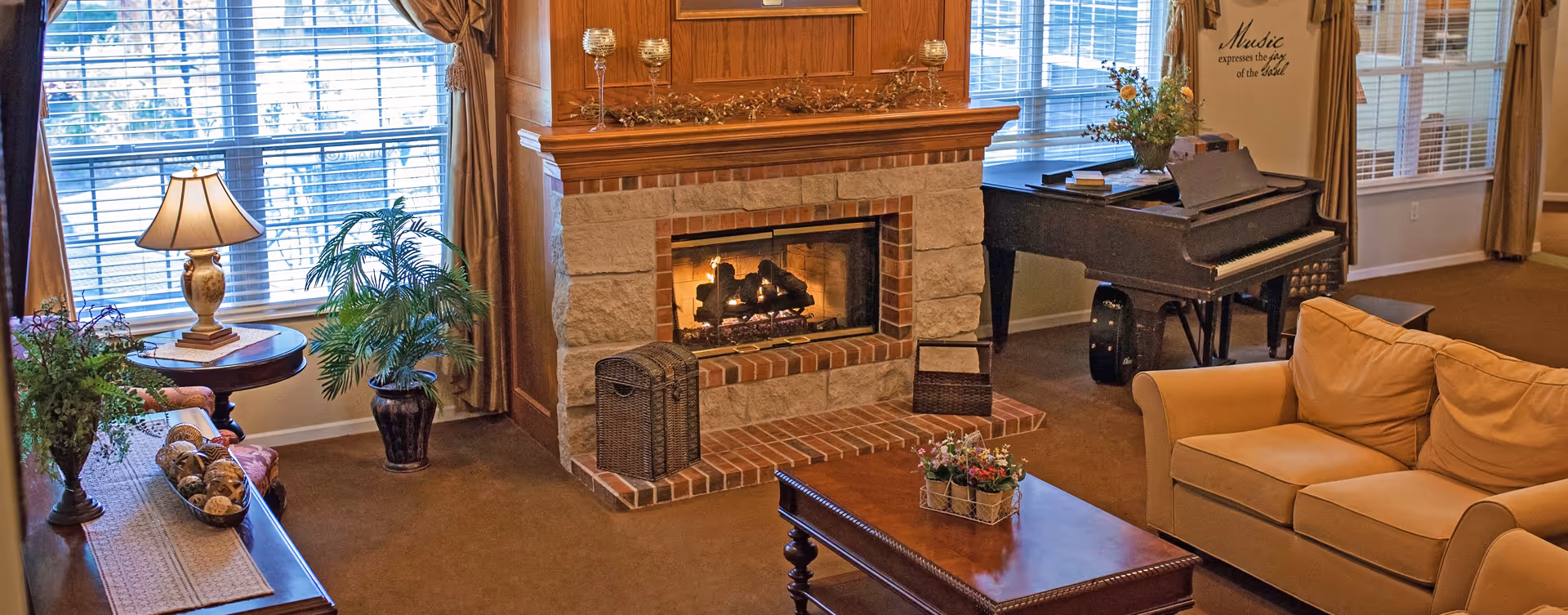 Cozy living room with a lit stone and brick fireplace, a beige sofa, a wooden coffee table with a small flower arrangement, a grand piano near the window, and decorative plants and lamps around the room.