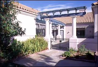 Outdoor view of a residential care facility entrance with a tiled roof, white walls, a black metal gate, and a pergola structure above the gate. There are plants and shrubs along the pathway leading to the entrance.