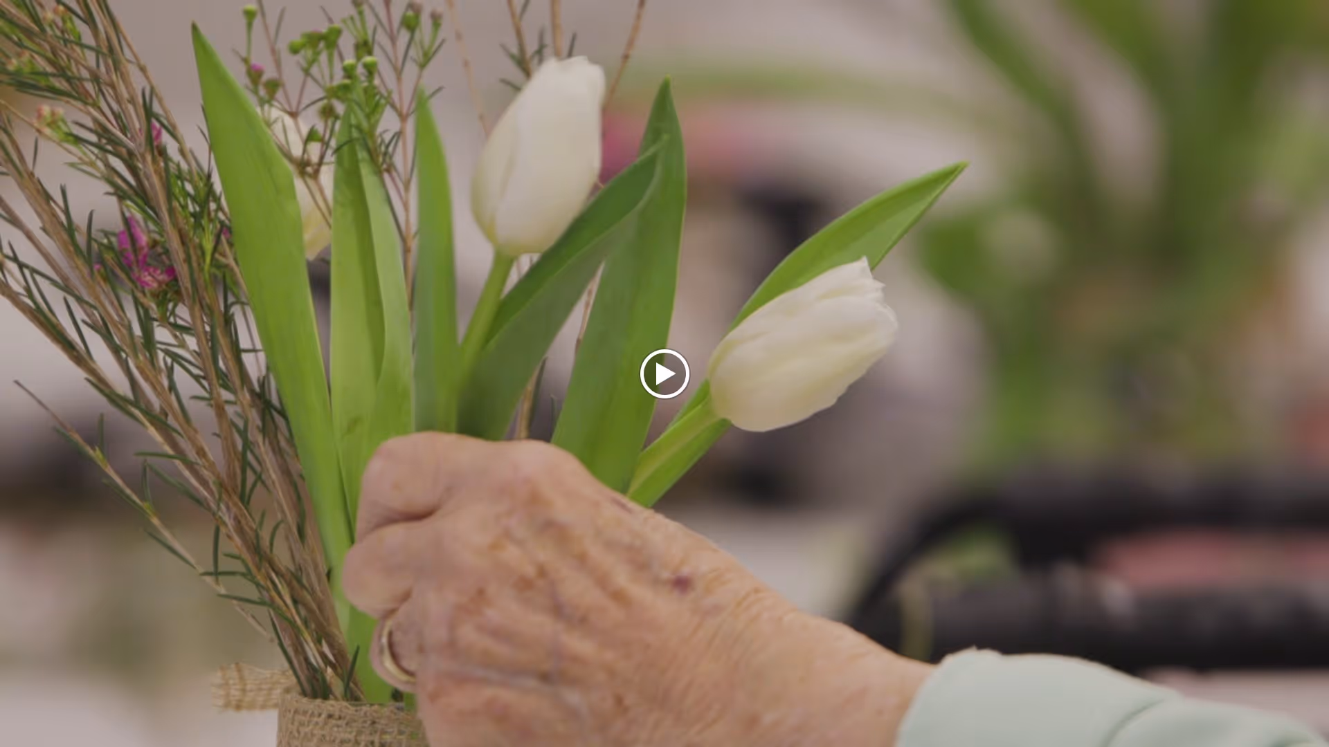 Close-up of an elderly person's hand arranging white tulips and greenery in a floral bouquet indoors.