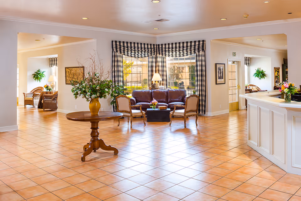 A spacious and well-lit common area in a senior living facility featuring a round wooden table with a large floral arrangement in a yellow vase in the center. In the background, there is a seating area with a brown leather sofa, two upholstered chairs, a coffee table with flowers, and large windows with black and white checkered curtains. To the right, there is a white reception desk with a flower vase. The floor is tiled with a warm terracotta color, and the walls are painted light beige. Additional seating with wicker chairs and plants is visible in adjacent rooms.