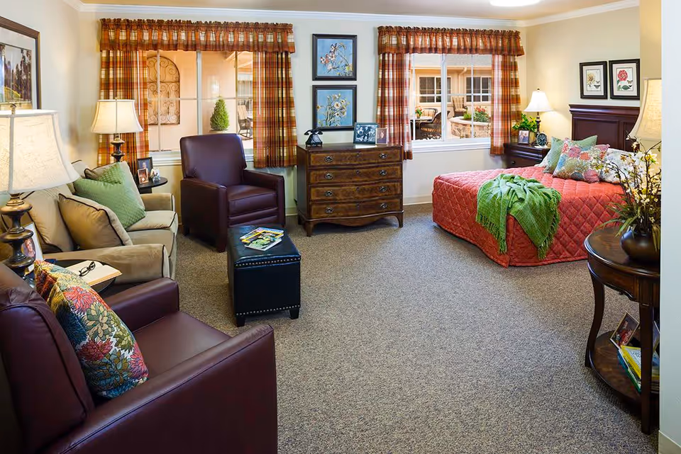 A cozy, well-lit bedroom in a senior living facility featuring a bed with a red quilt and green throw blanket, two windows with plaid curtains, a wooden dresser with framed photos and decor, two armchairs with colorful pillows, a beige sofa, several lamps, and a small round table with flowers and books.