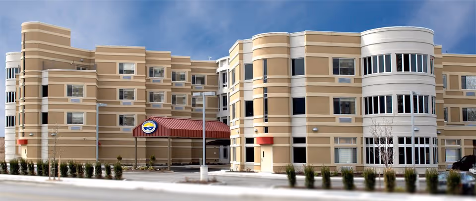 Exterior view of a multi-story beige and white rehabilitation and nursing care center building with numerous windows and a covered entrance with a red roof. The sky is clear and blue.